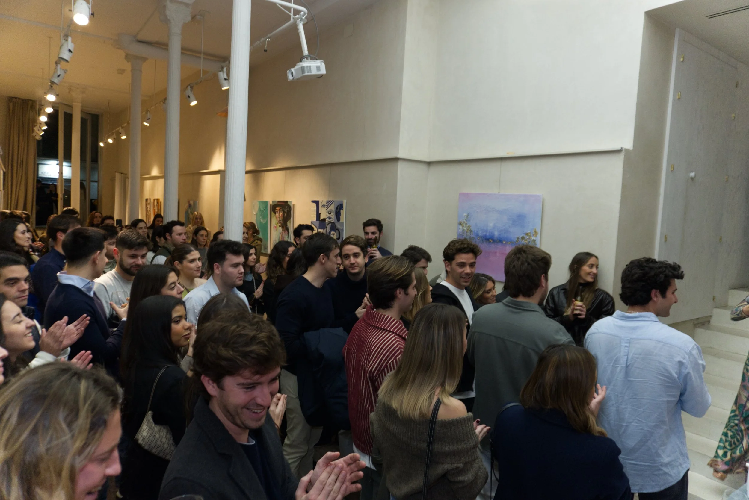 Un grupo de personas en una galería de arte, observando y aplaudiendo las obras en exhibición y algunas sonriendo.