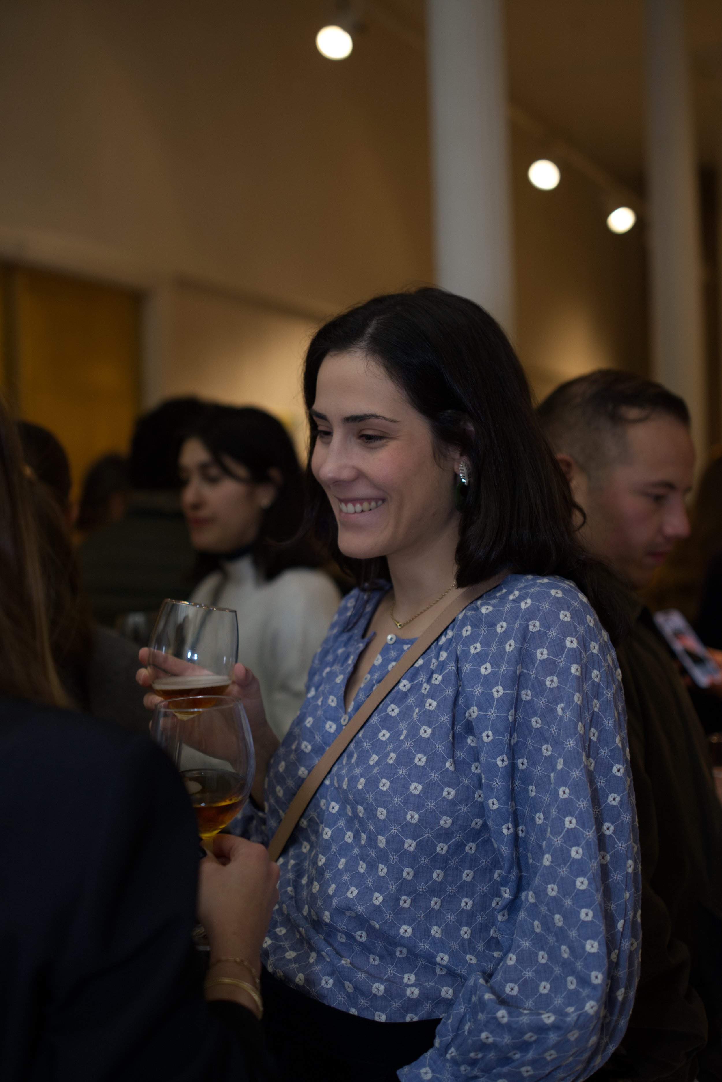 Una mujer sonriendo sostiene una copa de vino en un evento social, rodeada de otras personas en un ambiente elegante y acogedor.