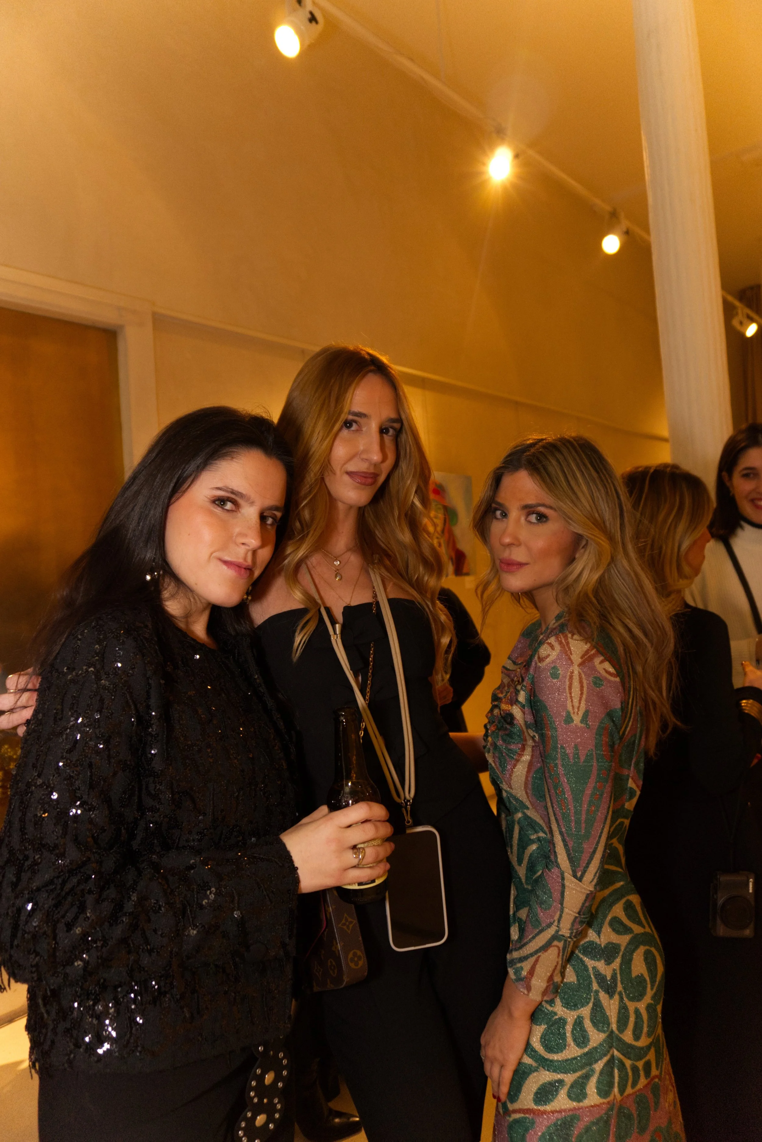 Mujeres en una fiesta elegante, posando para la foto, una de ellas sostiene una botella y llevan prendas formales.