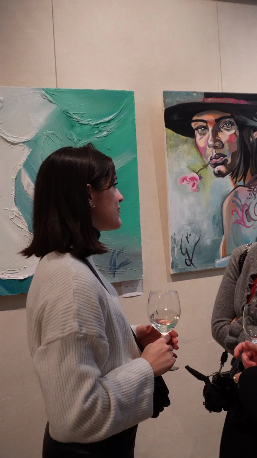 Mujer con cabello castaño oscuro, sosteniendo una copa de vino, de perfil, en una galería de arte con pinturas en la pared.