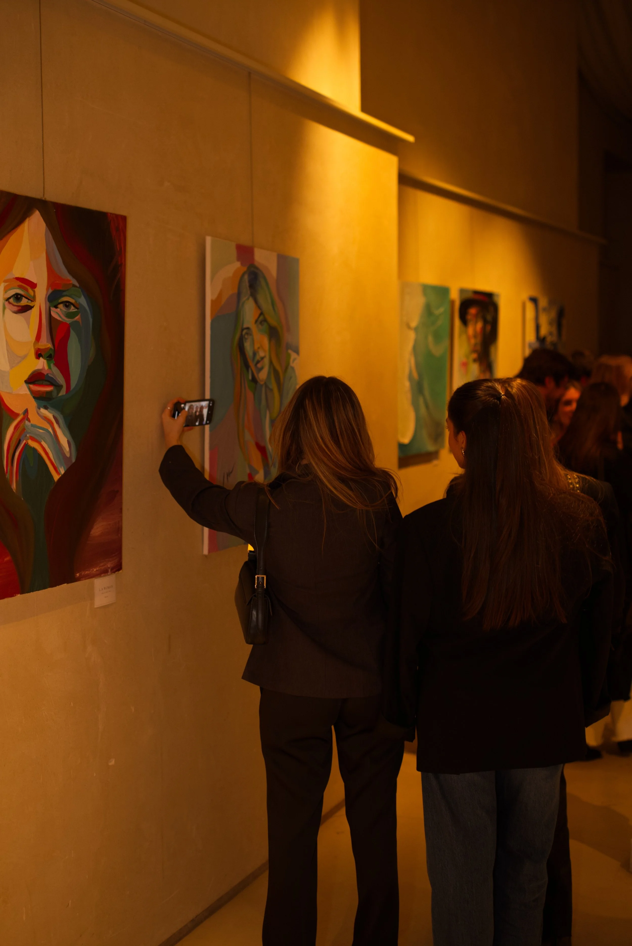 Personas observando pinturas coloridas en una exposición de arte, algunas tomando fotos.
