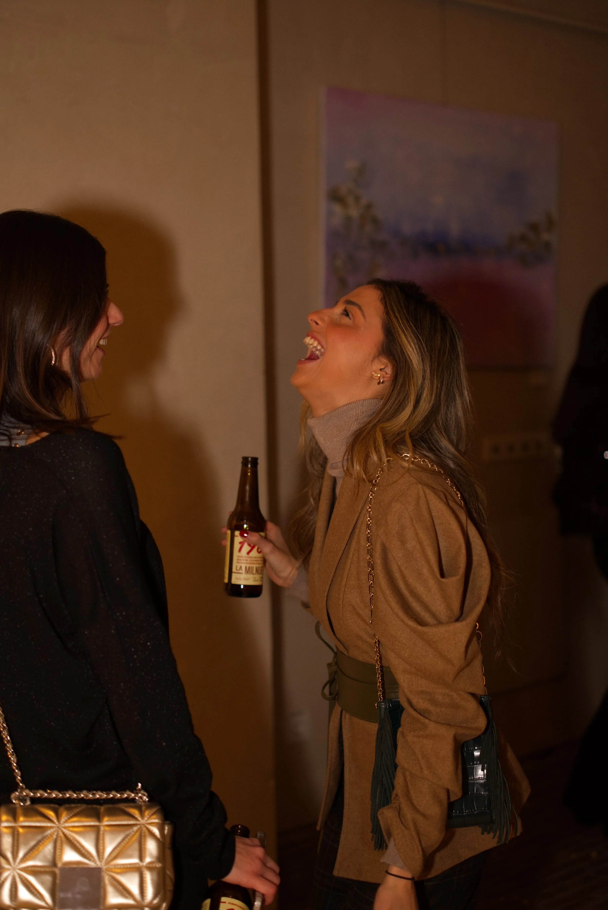 Dos mujeres riendo y conversando en una reunión social, una de ellas sostiene una botella de cerveza.