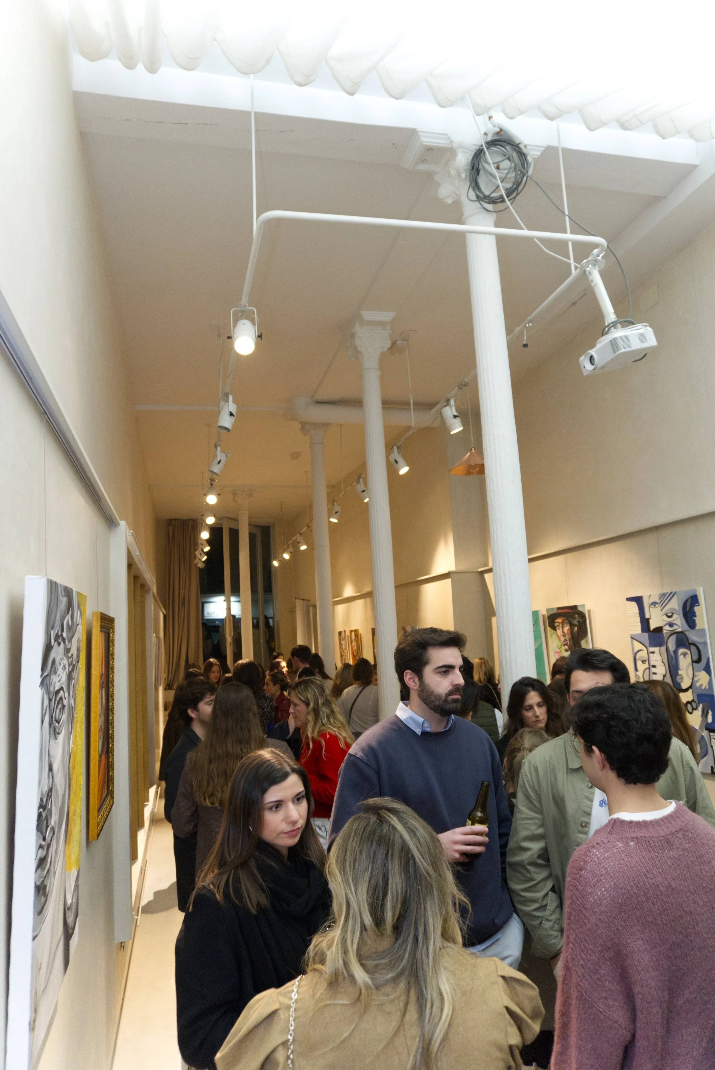 Galería de arte con muchas personas observando pinturas en las paredes, ambiente iluminado con luz de spot y decorado con columnas blancas.