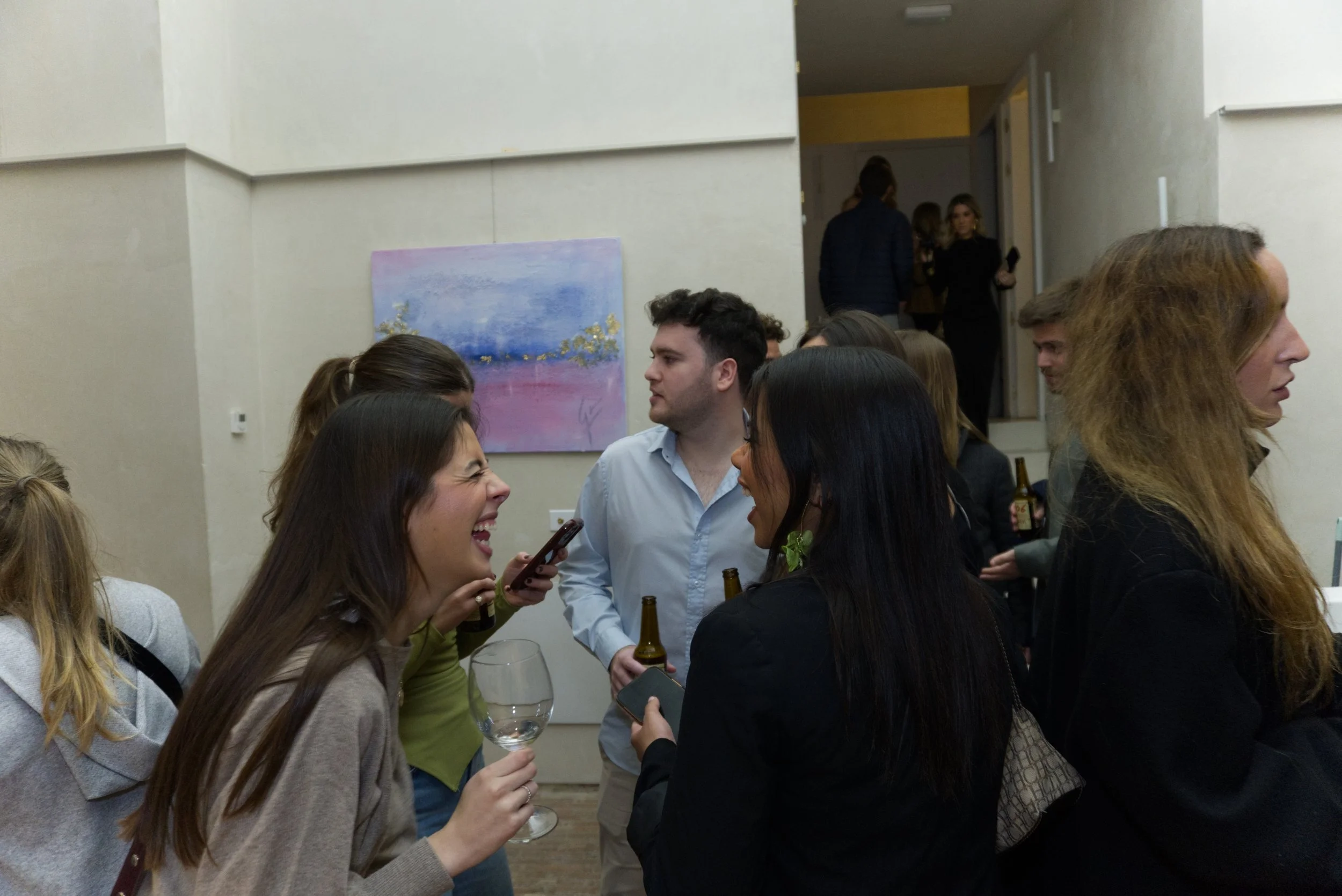 Personas socializando en una galería de arte, algunas con bebidas y teléfonos, en un ambiente de interiores con pintura en la pared.