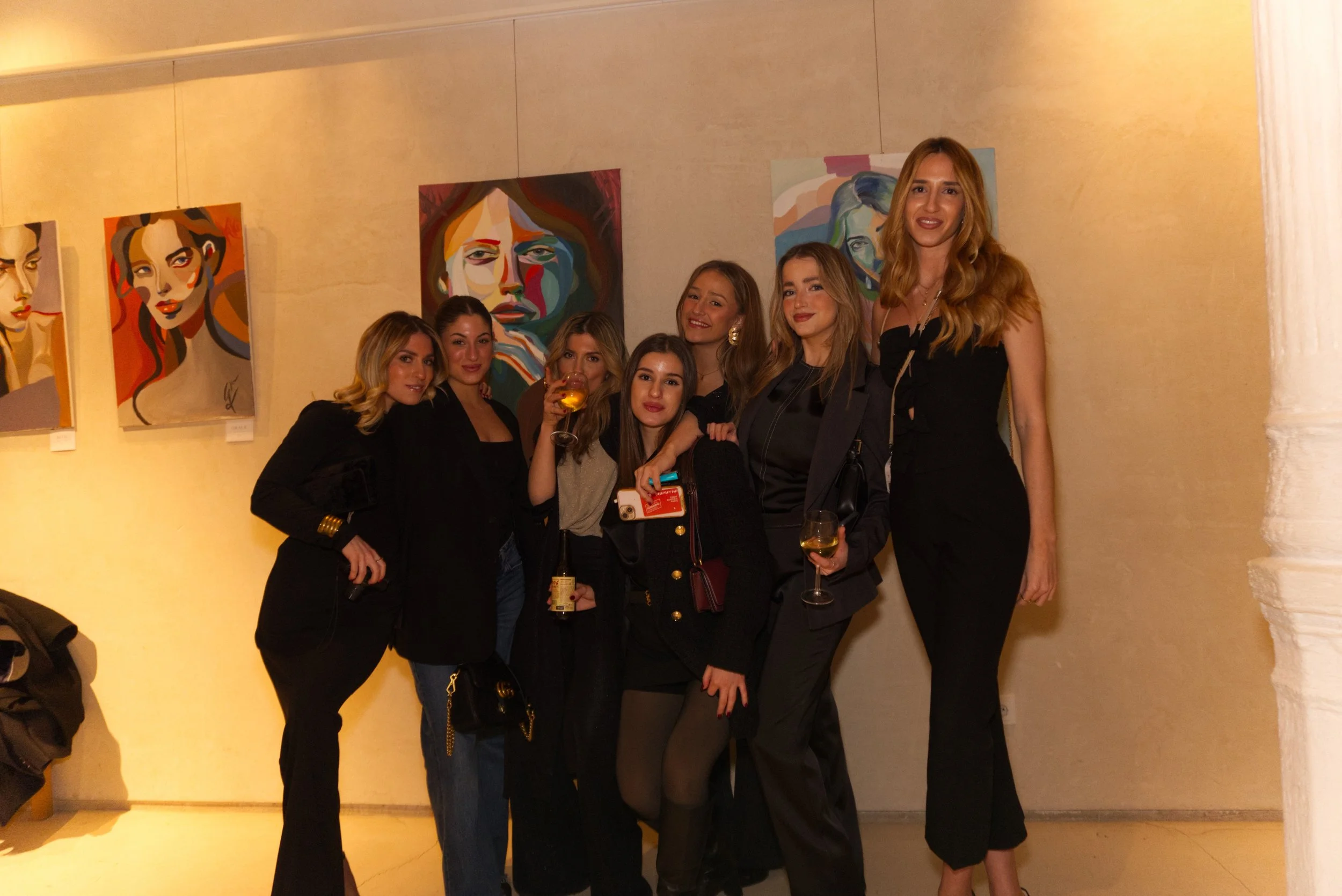 Grupo de siete mujeres jóvenes en una galería de arte, posando junto a pinturas de retratos abstractos, en un evento social con pintura y vino.