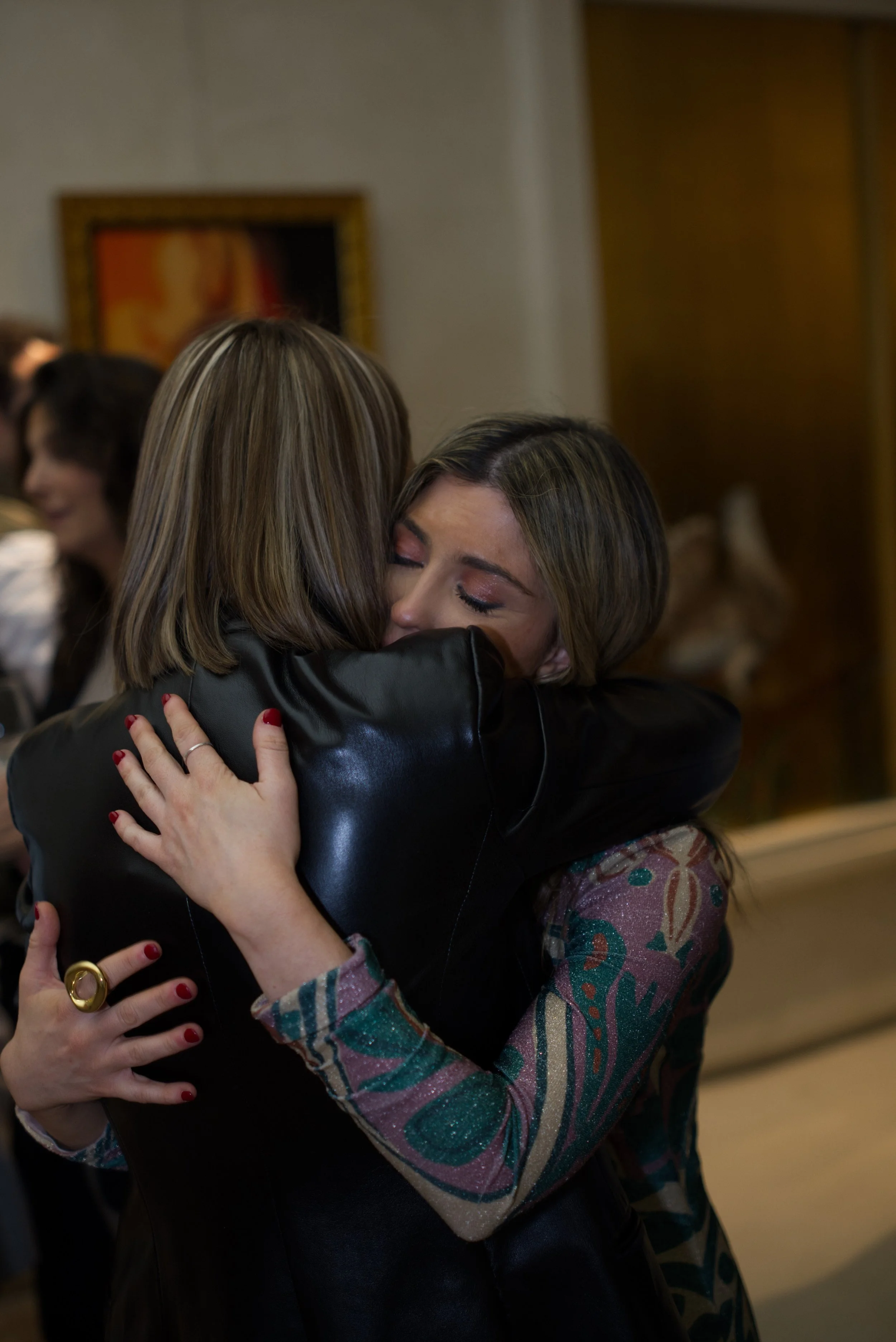 Dos mujeres abrazándose con emoción en un ambiente social, con otras personas en el fondo.