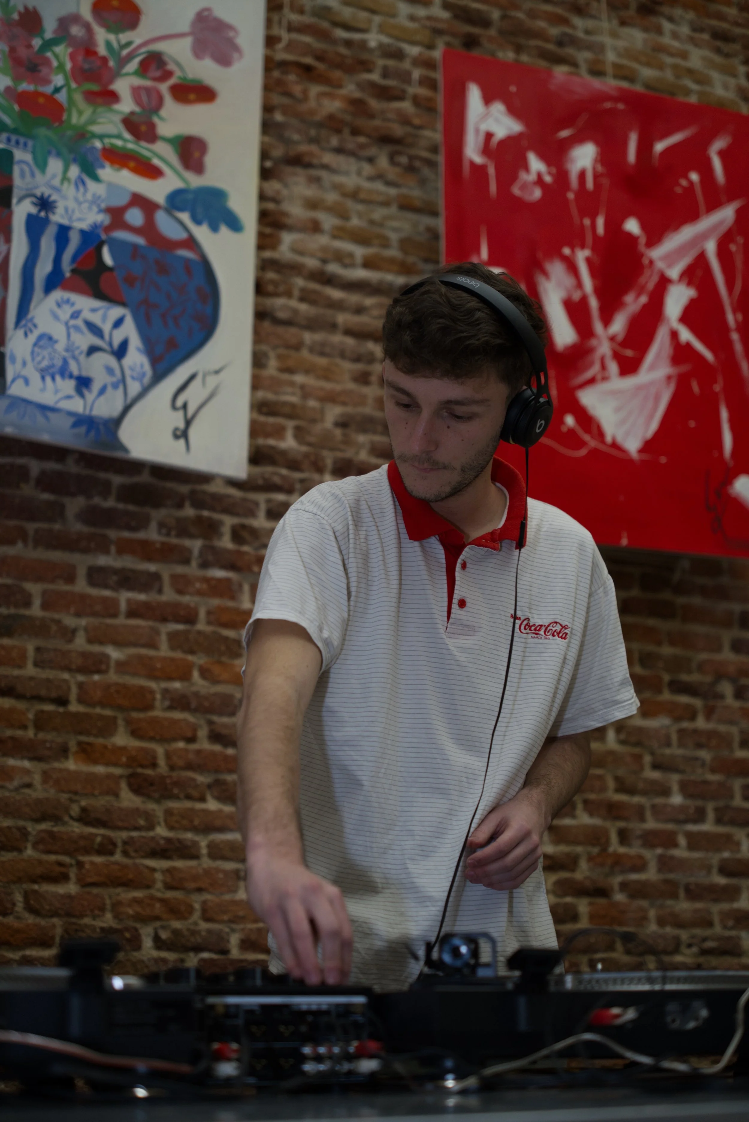 Joven DJ con auriculares mezclando música en un estudio con paredes de ladrillo, decorado con arte abstracto en color rojo y una botella decorativa con flores en el fondo.