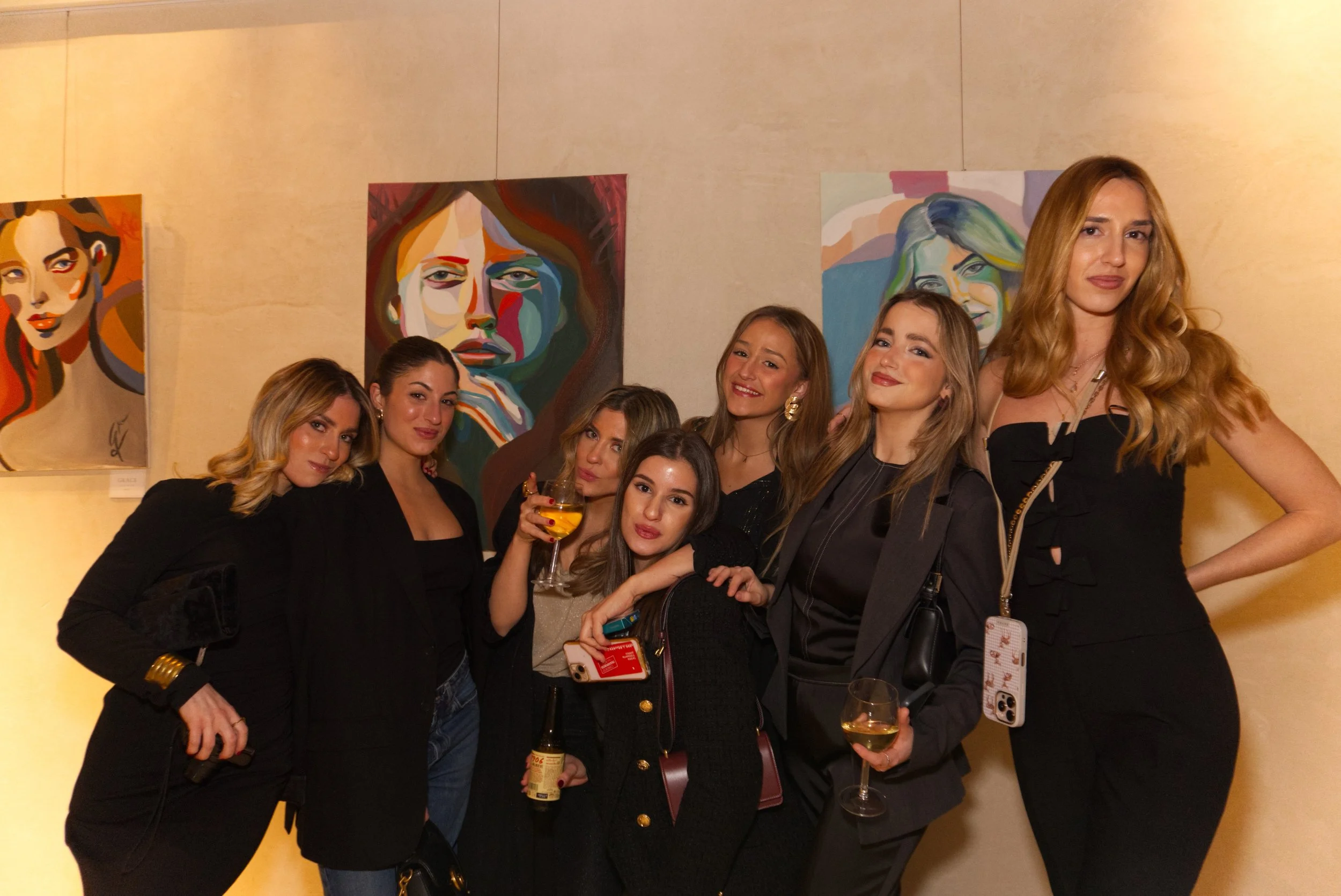 Grupo de siete mujeres posando en una galería de arte con retratos coloridos en el fondo, algunas sosteniendo bebidas y con teléfonos en mano.