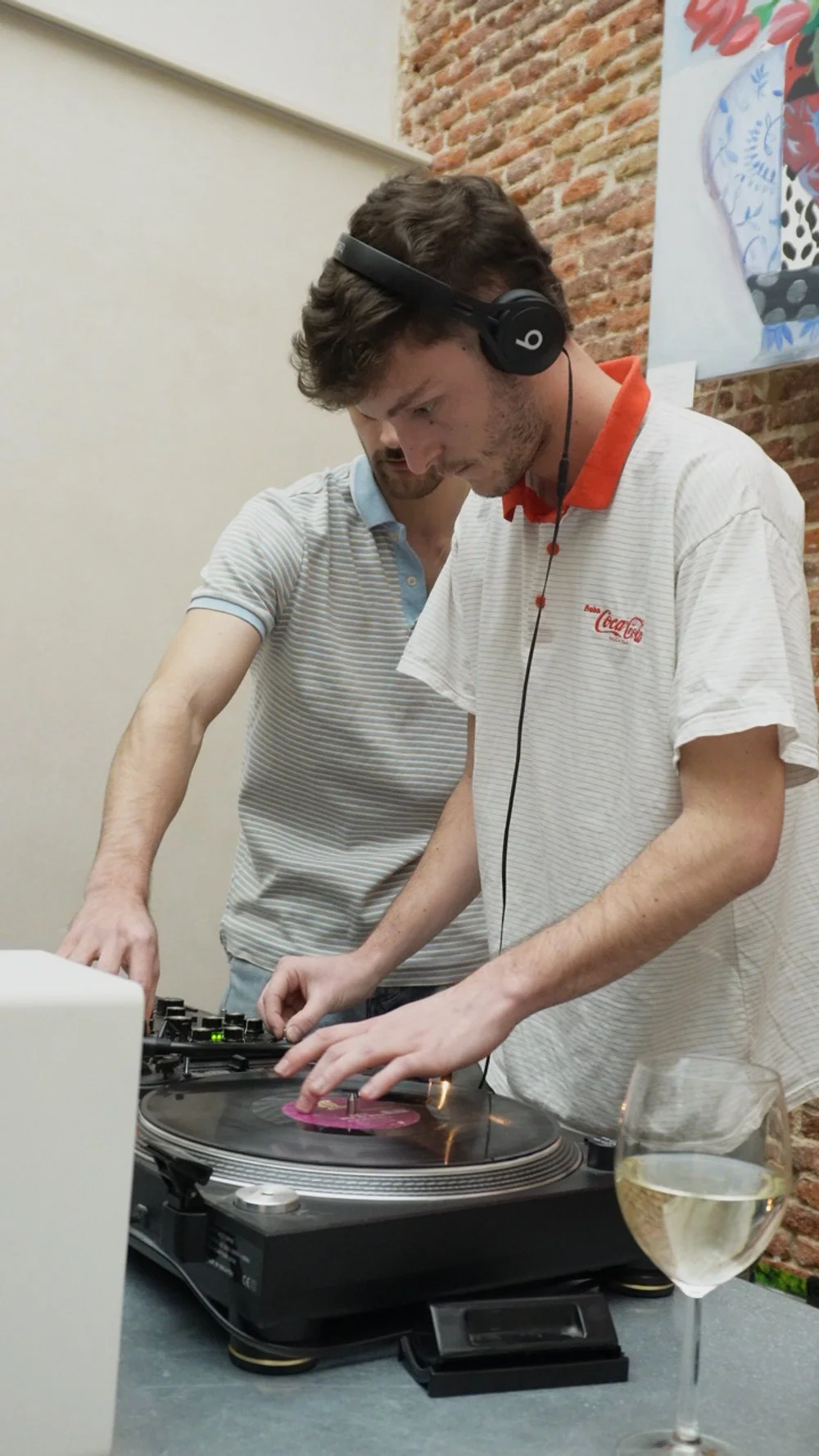 Dos jóvenes mezclando música con un tocadiscos en una mesa, con una copa de vino al frente.