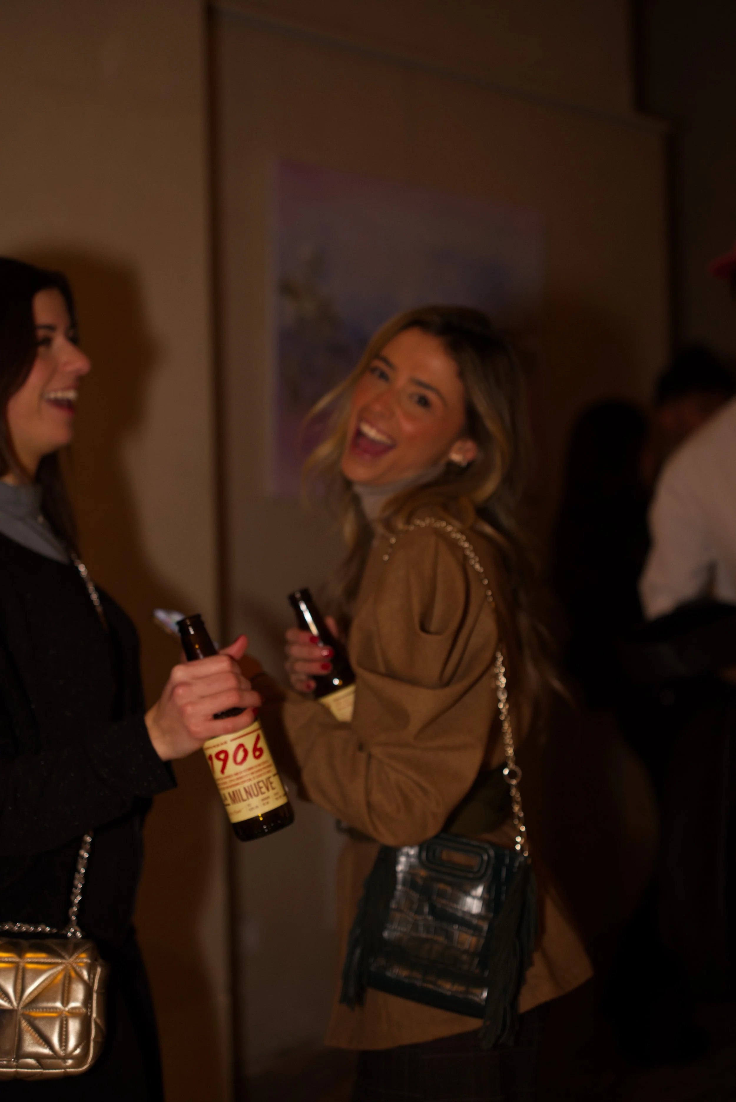 Dos mujeres en una fiesta, una con una botella de cerveza y sonriendo mientras la otra conversa.