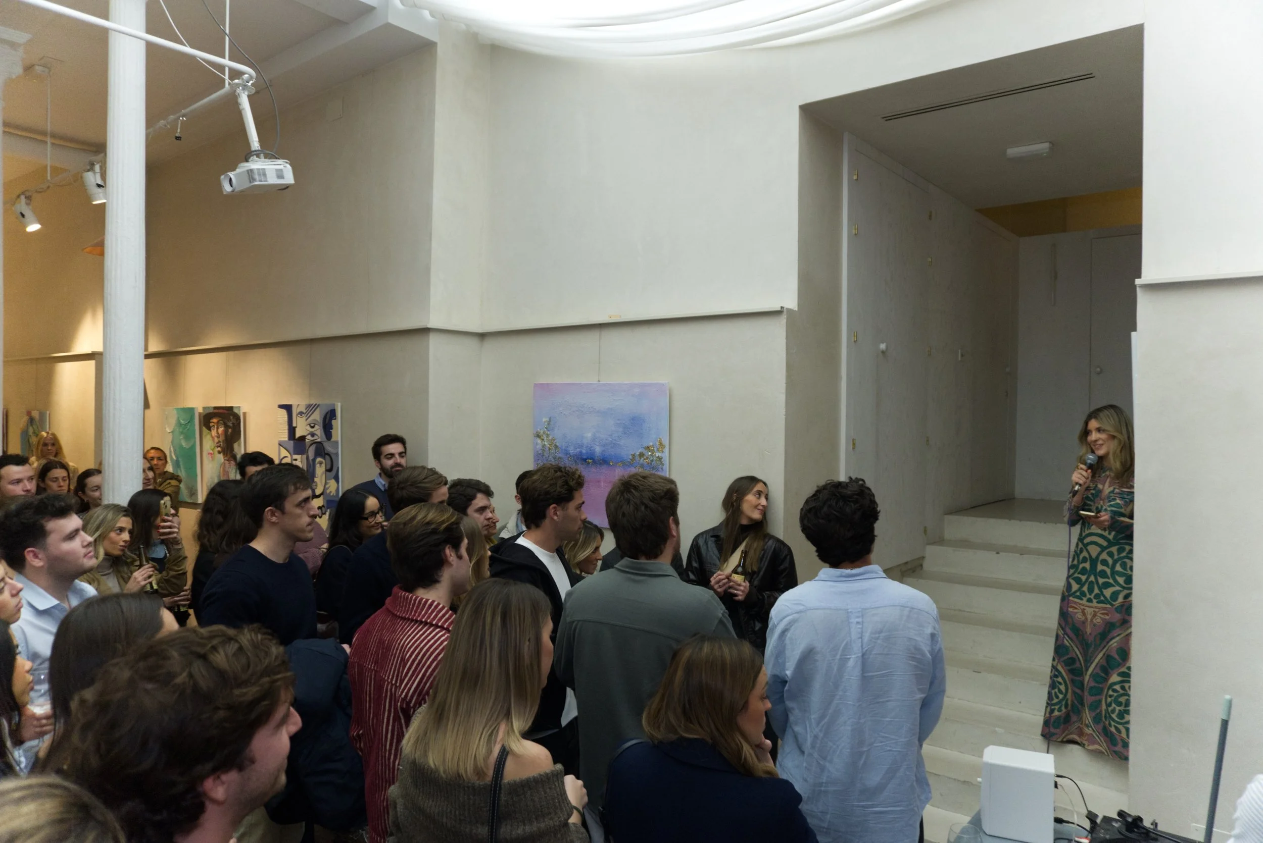 Una mujer con vestido largo de colores y estampados hablando con micrófono frente a un grupo de personas en un espacio de arte, con pinturas en las paredes.