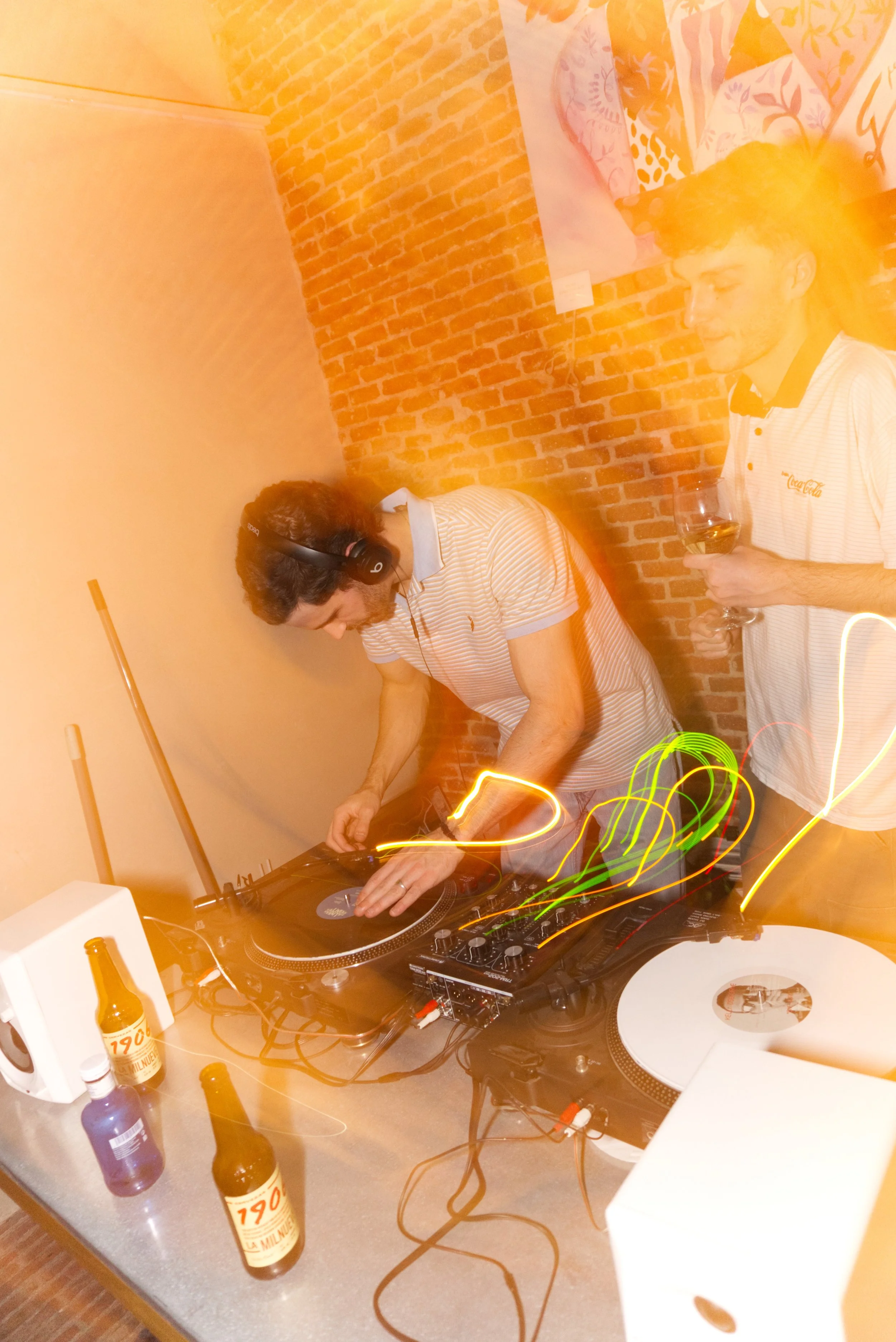 Dos hombres en una fiesta, uno es DJ mezclando música con auriculares y el otro está sosteniendo una copa de vino, en un ambiente con paredes de ladrillo y decoraciones coloridas.
