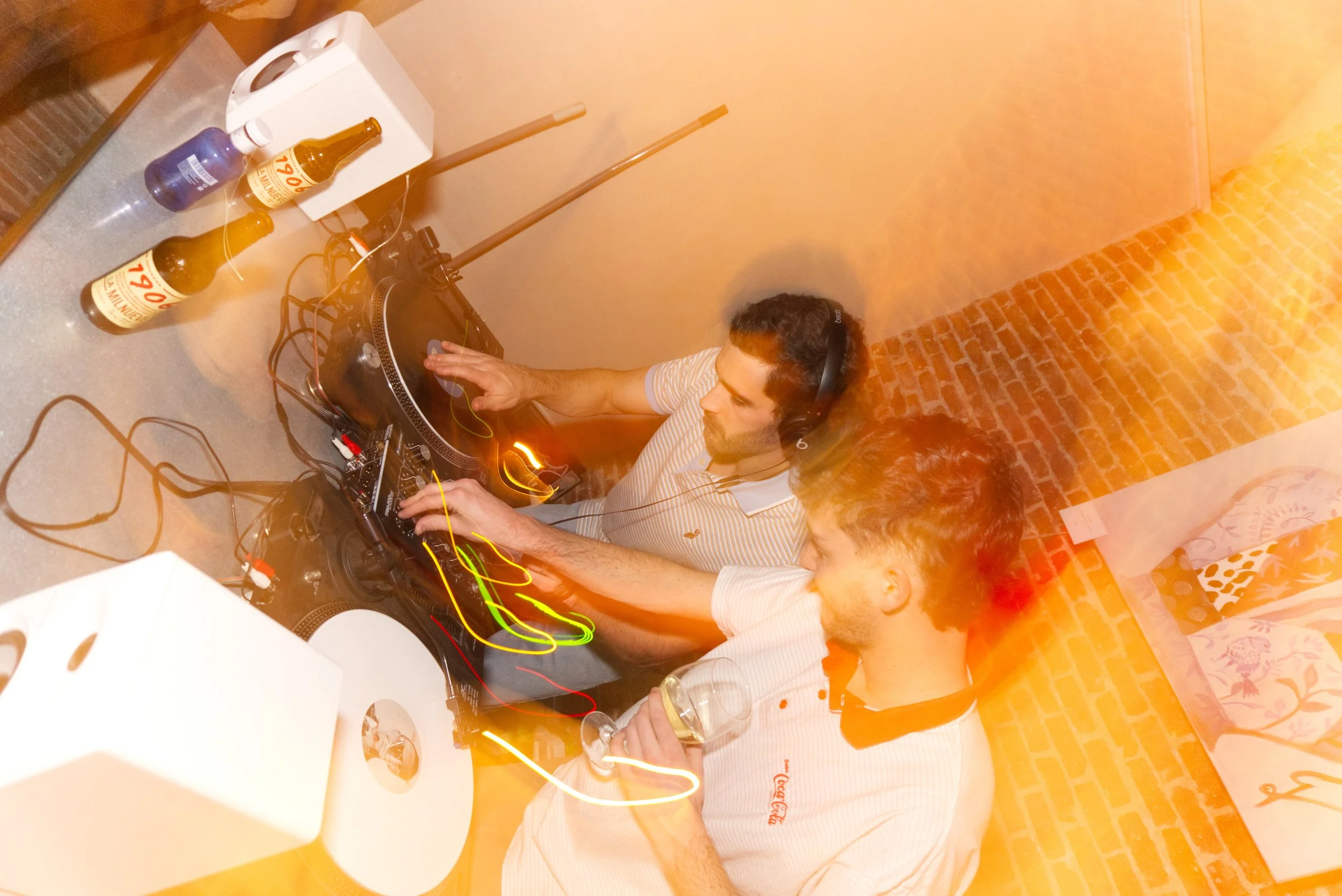Dos hombres DJs mezclando música en un set en un ambiente interior con pared de ladrillo y decoraciones de tela con estampados