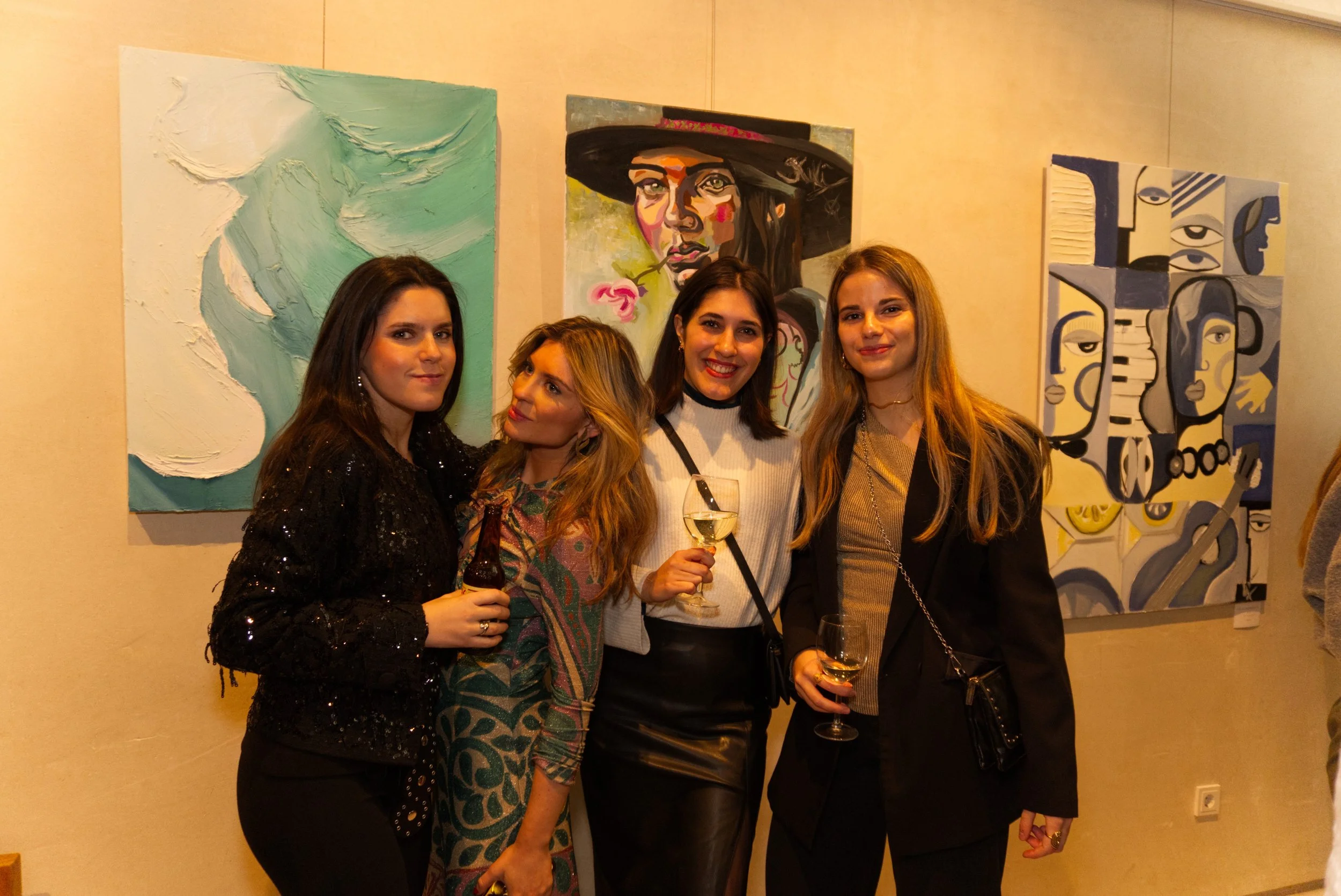 Cuatro mujeres en una galería de arte, sosteniendo bebidas, con pinturas abstractas de colores vivos en el fondo.
