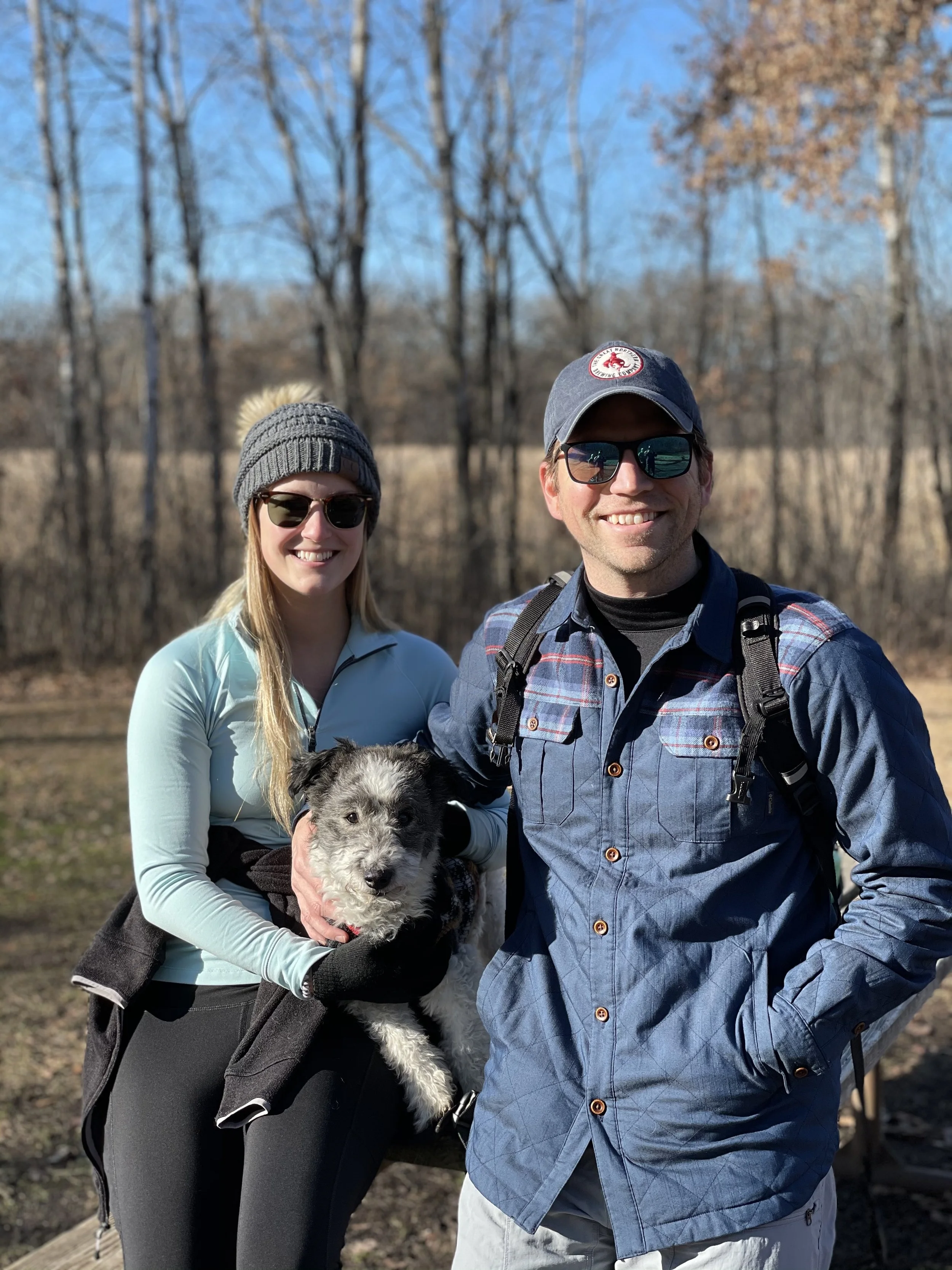 A smiling young woman and man outdoors in fall or winter with a dog. The woman is holding the dog, wearing sunglasses, a gray knit beanie, and a light blue jacket. The man is wearing sunglasses, a gray cap, a blue checkered shirt, and has a backpack.