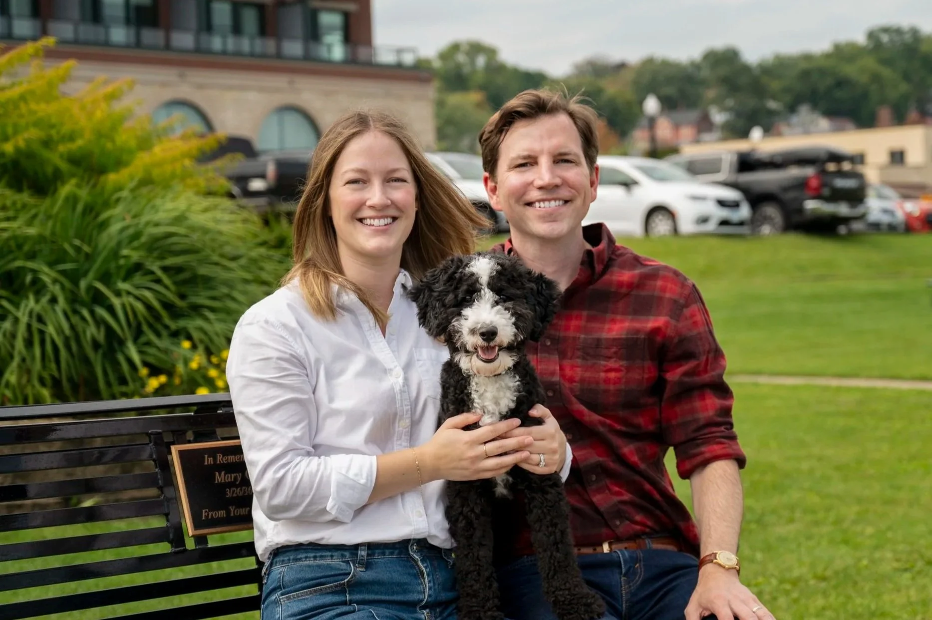 A smiling couple sitting on a park bench holding a black and white puppy, with greenery and parked cars in the background.