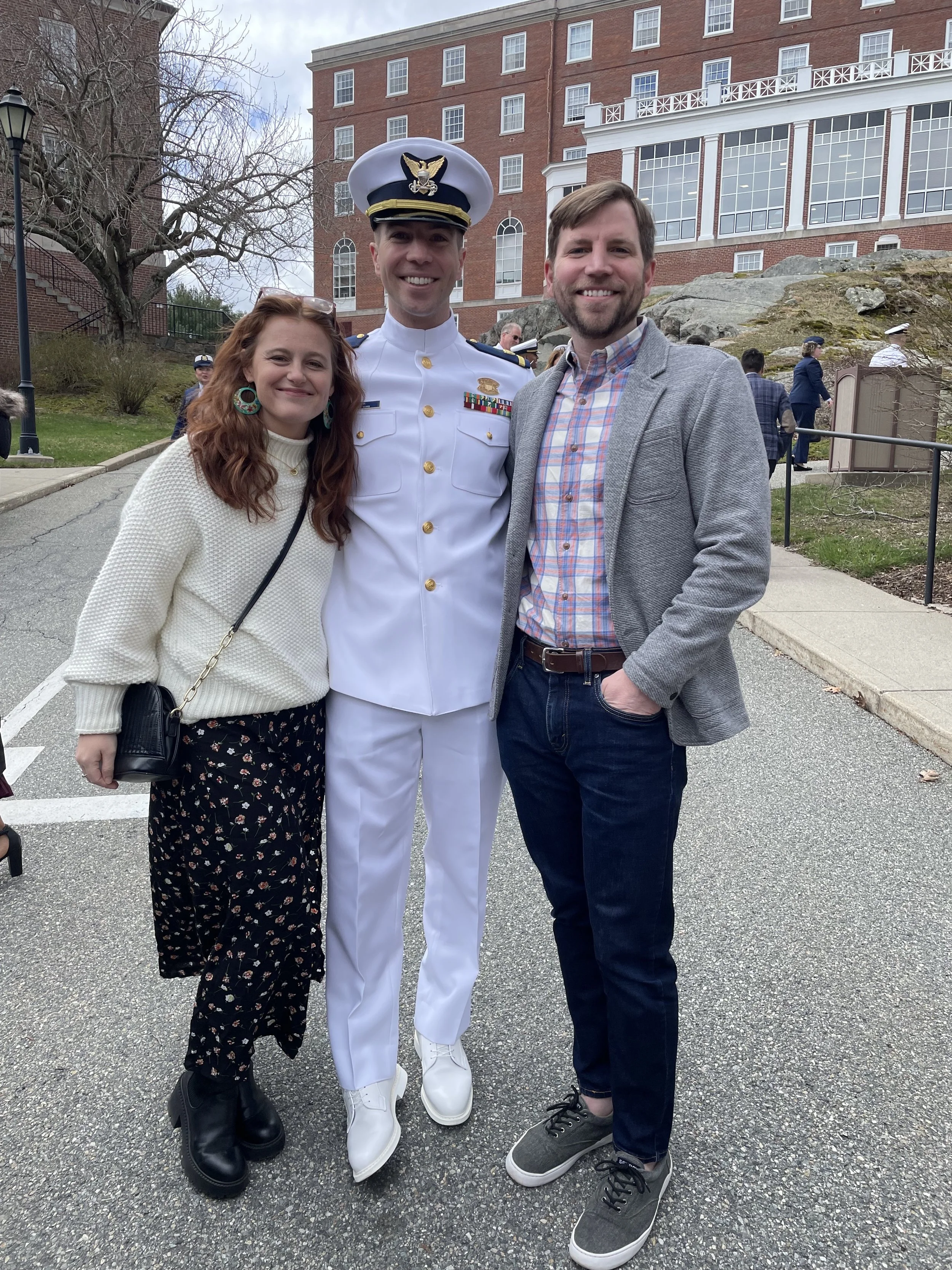 Three people standing together outside, smiling: a woman with long red hair in a white sweater and black floral pants, a man in a light gray blazer and dark jeans, and a man in a white military dress uniform with a white hat, decorated with medals. T