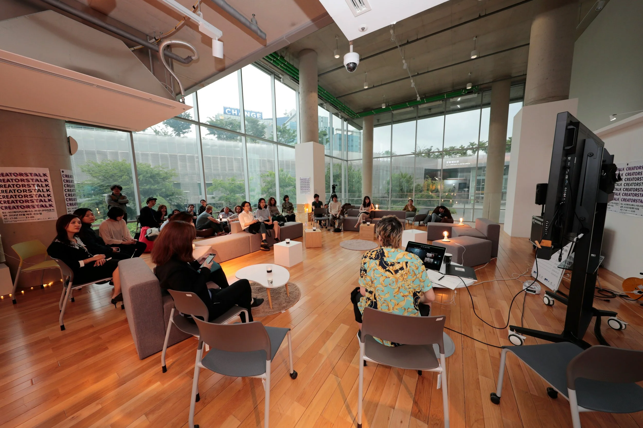 A group of people attending a seminar or workshop in a modern, spacious room with large glass windows and wood floors. The presenter sits at a table with a laptop, facing the audience.