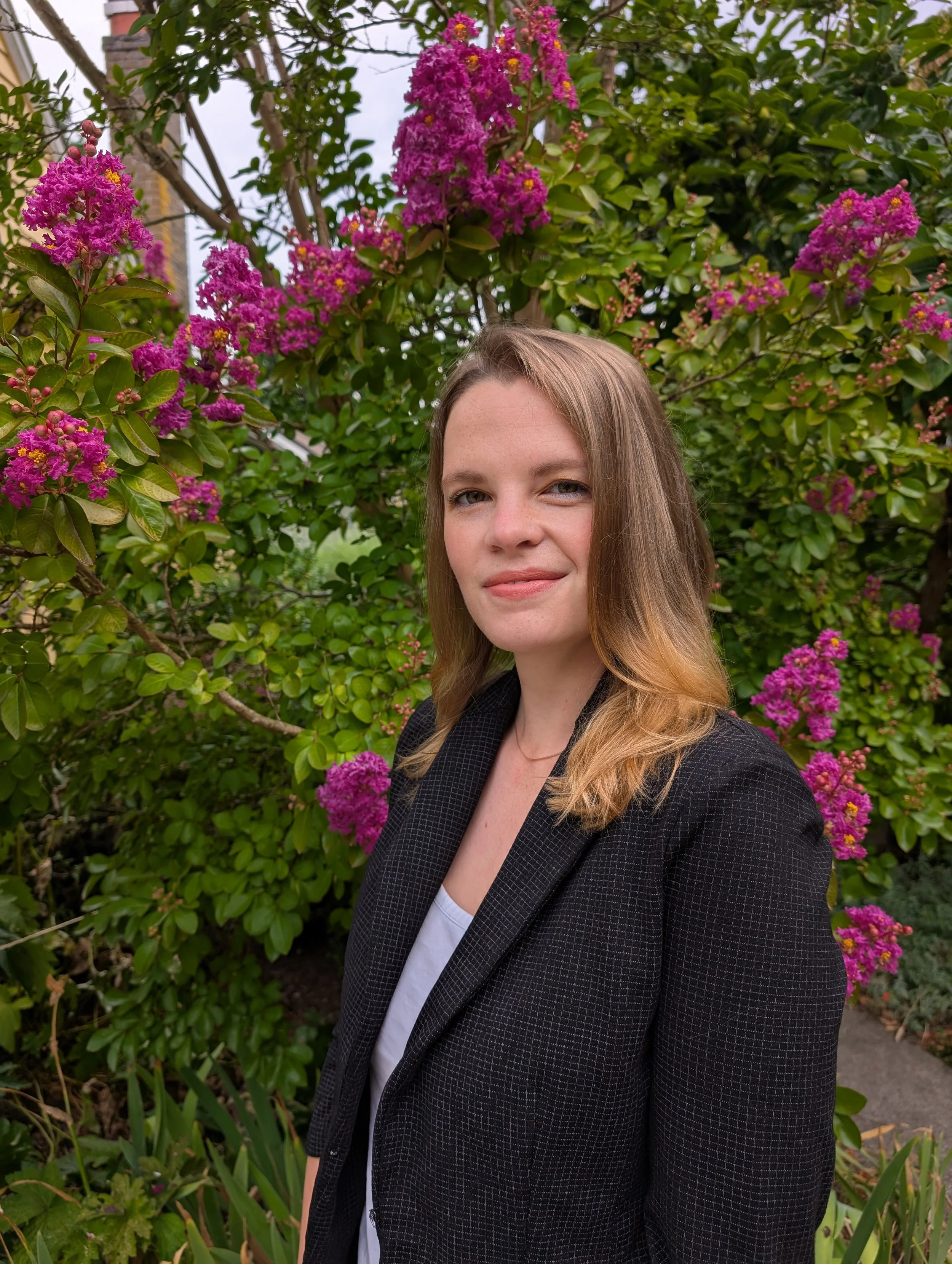 A woman with long light brown hair and a slight smile standing in front of a lush bush with pink and purple flowers.