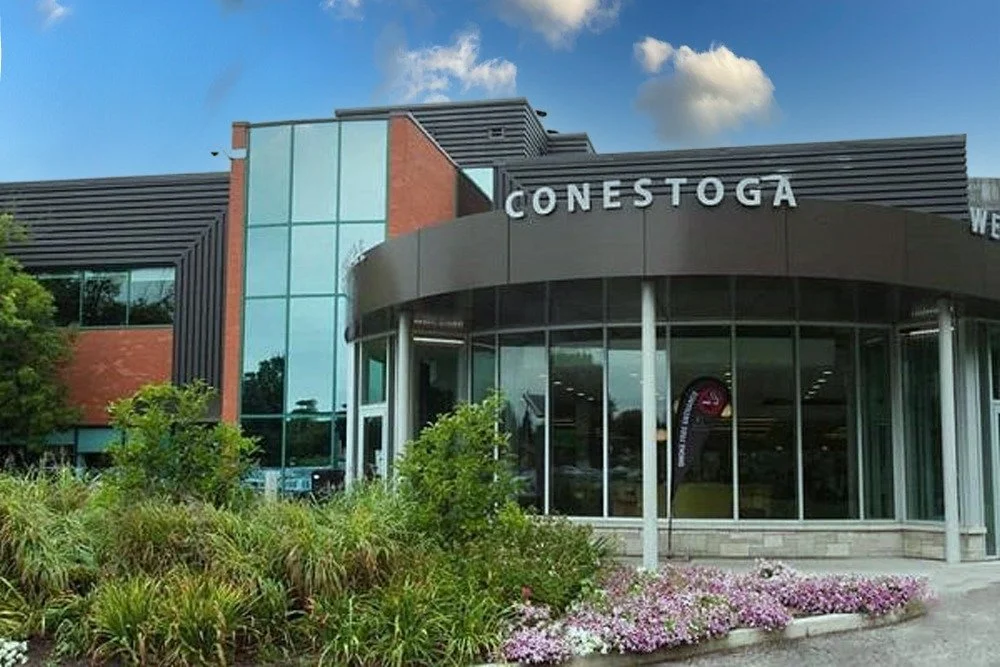 Exterior of a modern building with large glass windows and the sign 'CONESTOGA' at the entrance, surrounded by landscaped greenery and flowers.