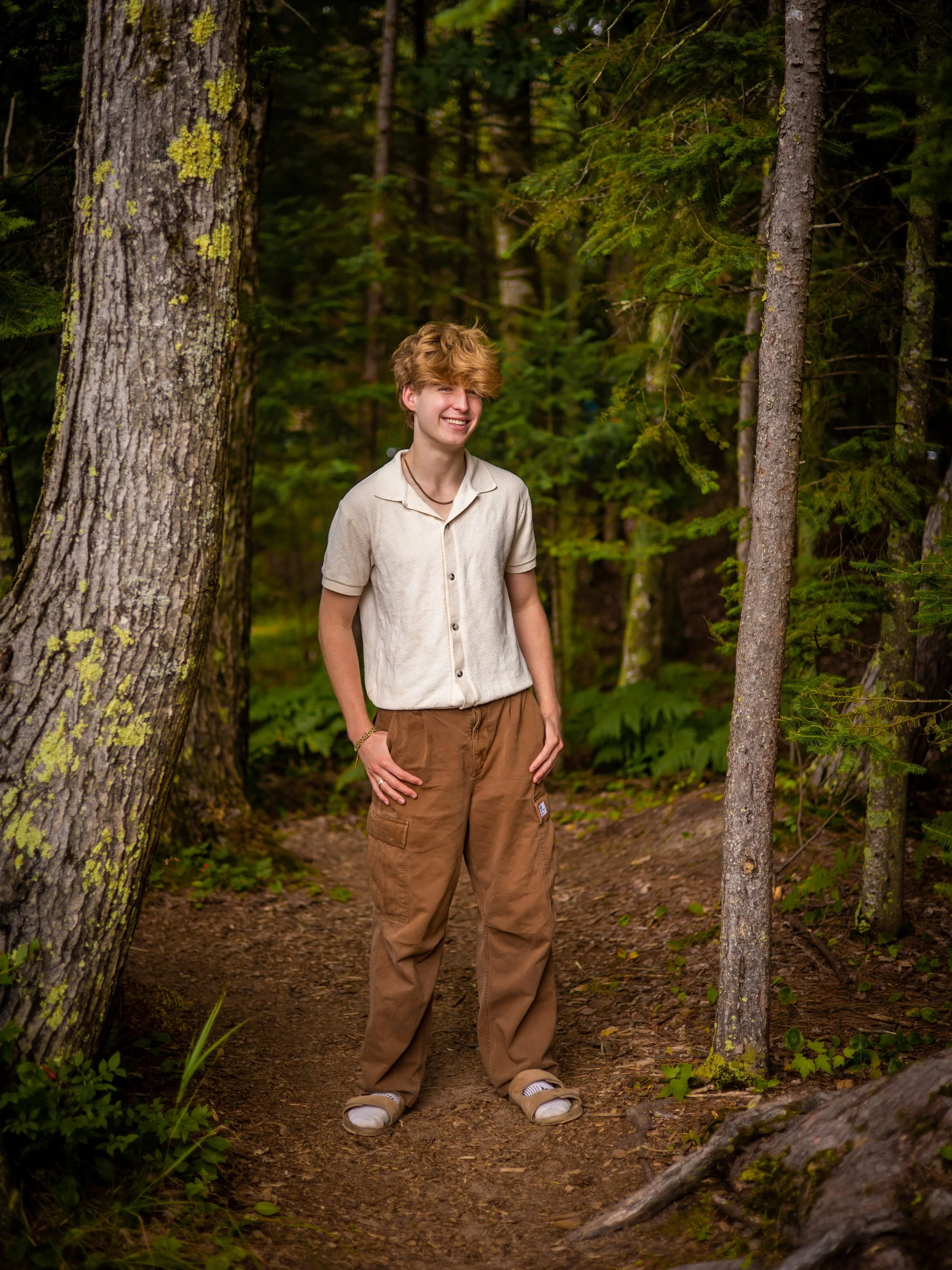 A young man with curly blond hair smiling and standing on a forest trail, surrounded by tall green trees and lush foliage.