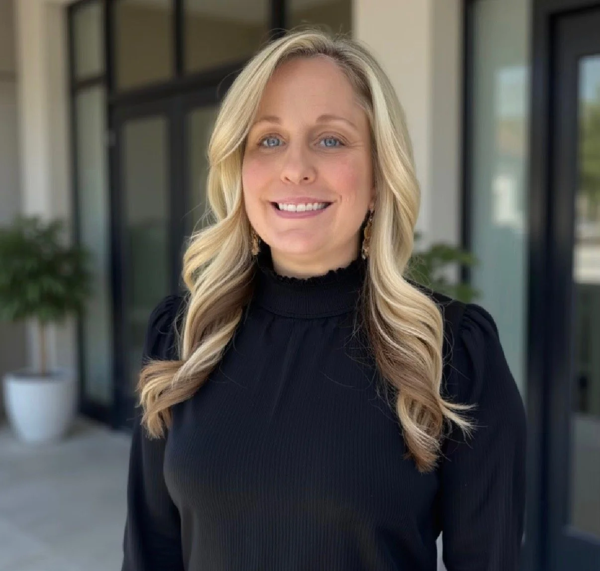 Smiling blonde woman in a black blouse standing indoors near glass doors