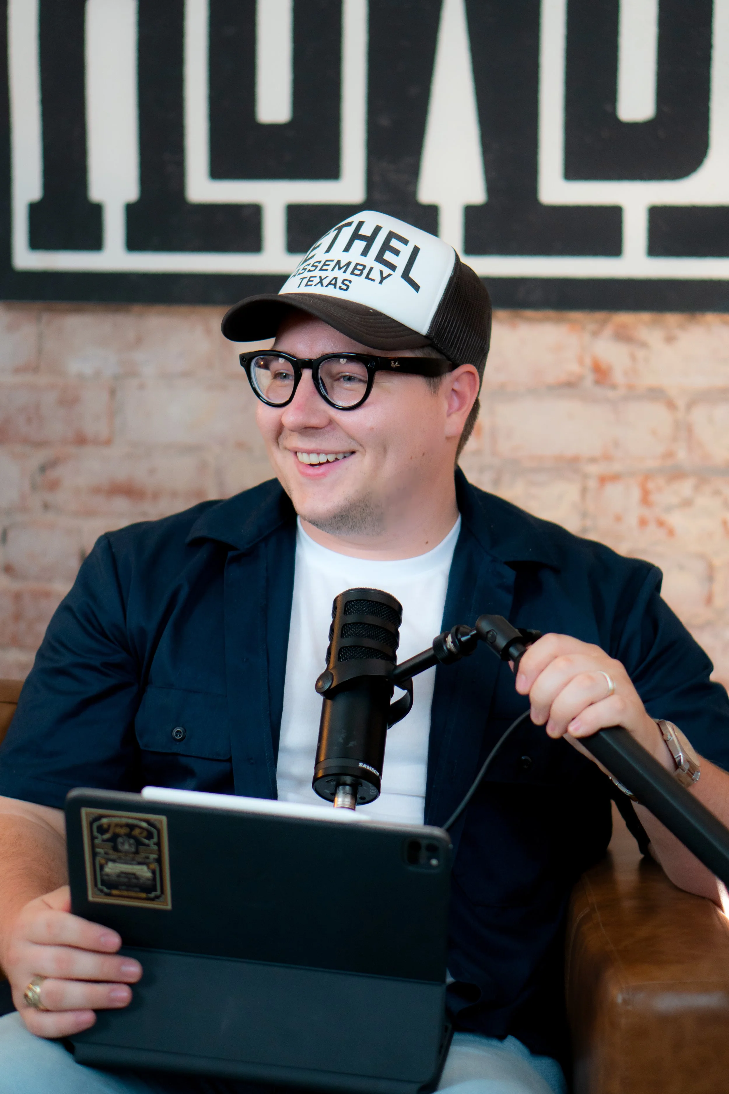 Smiling man wearing glasses, a cap that says 'Bethel Assembly Texas,' and a dark shirt, sitting at a table with a tablet and microphone in front of him against a brick wall background. Looks like a podcaster who teaches about content.