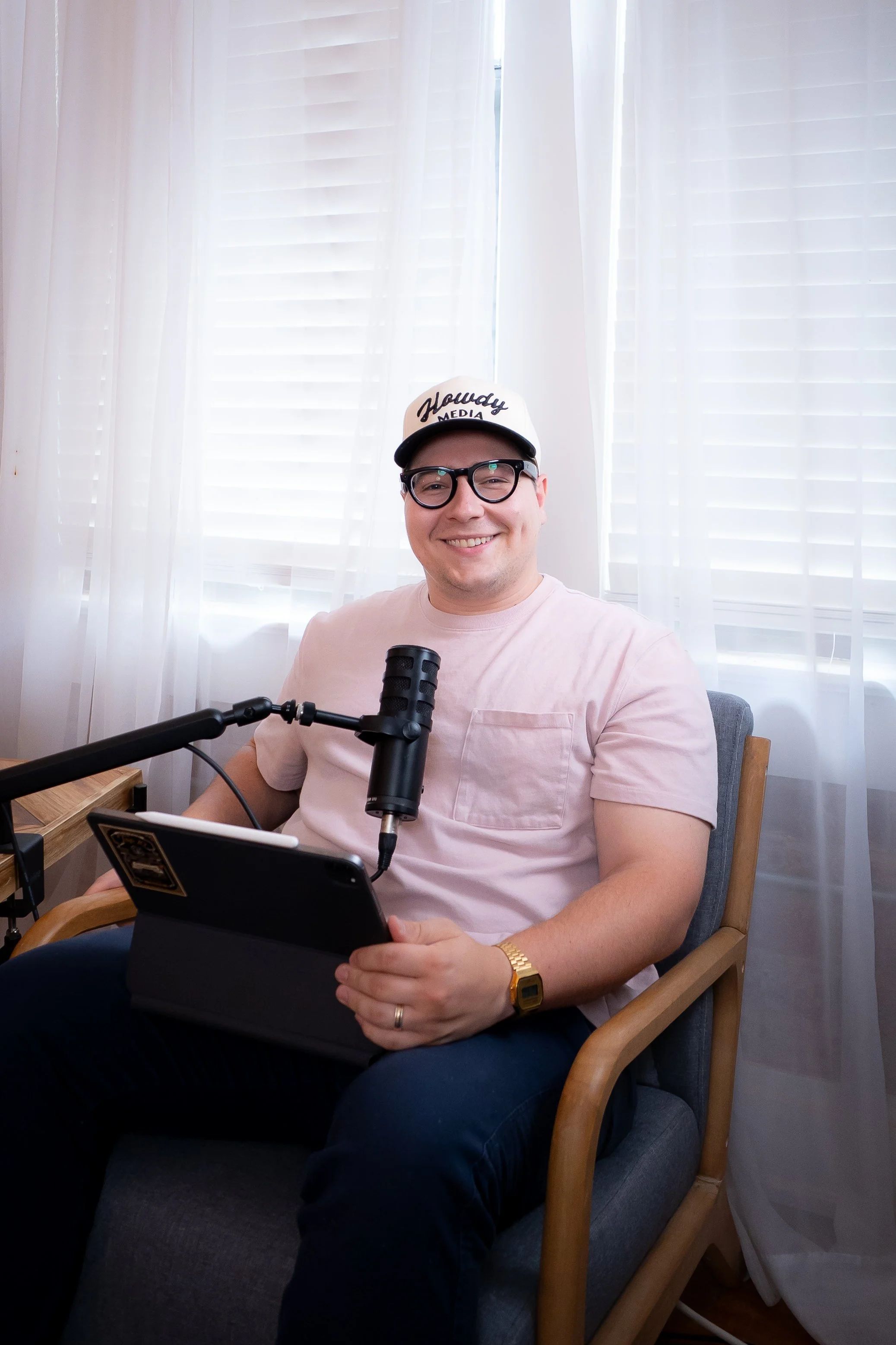 A man sitting in a chair with a microphone in front of him, holding a tablet, smiling, wearing glasses, a white cap, a pink t-shirt, and a gold watch, with white curtains and window blinds in the background.