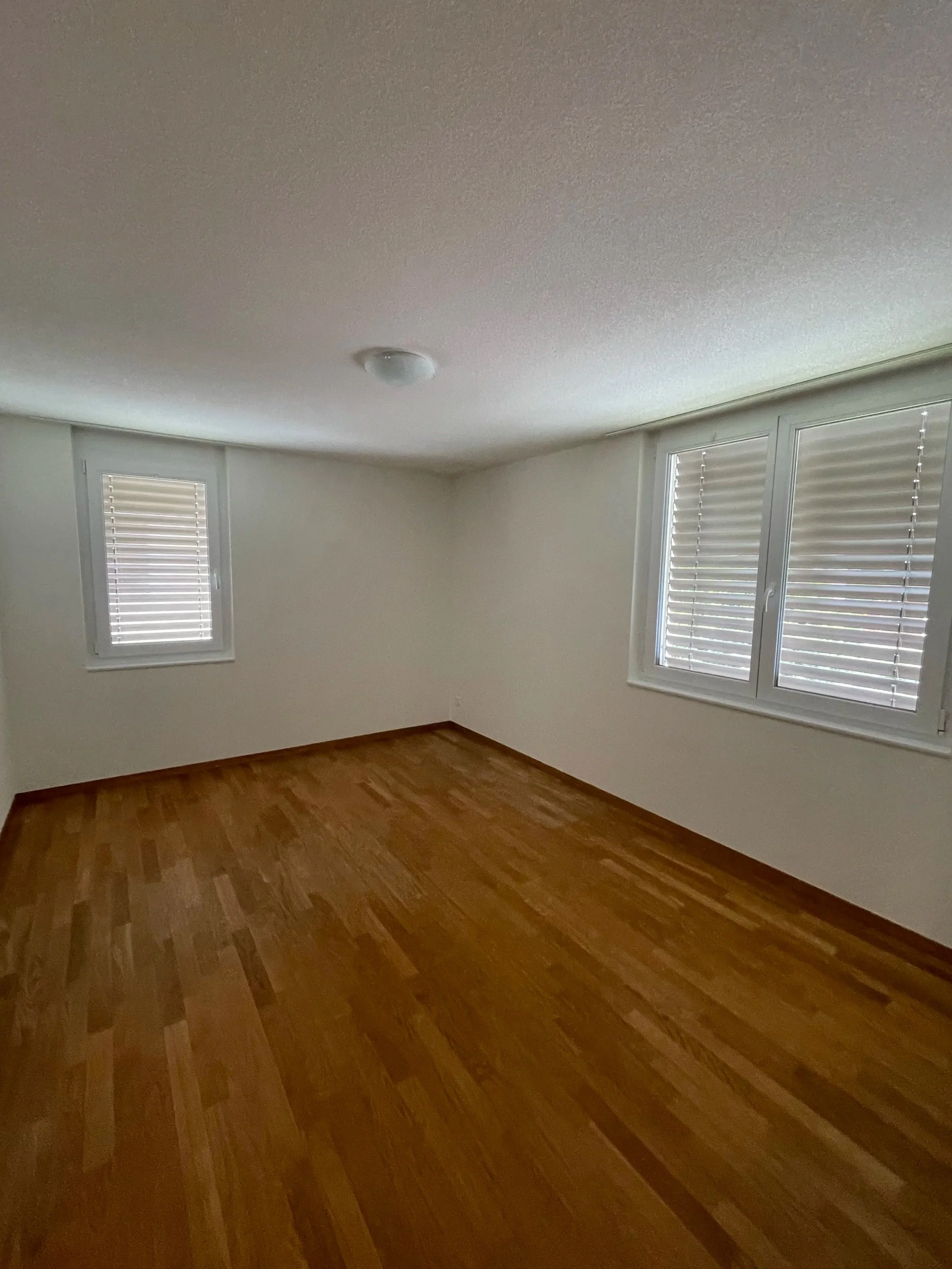 Ein leeres Zimmer mit weißen Wänden, zwei Fenster mit Rollläden und Holzboden.