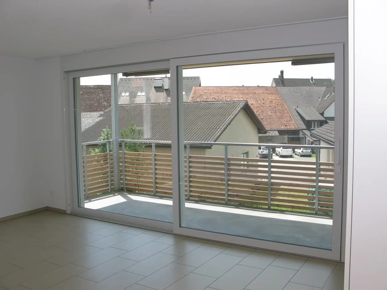 Innenraum eines Zimmers mit großen Fenster- und Türfronten, Blick auf ein kleines Balkongeländer und mehrere Häuser mit unterschiedlichen Dächern im Hintergrund.