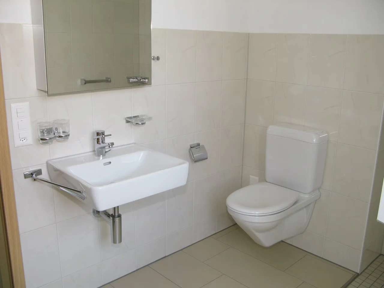 Kleines Badezimmer mit weißen Fliesen, Waschbecken, Spiegel, Spender für Seife oder Handtücher, Toilettenschüssel, Toilettepapierhalter, Wasserhahn, klein, sauber, schlicht.