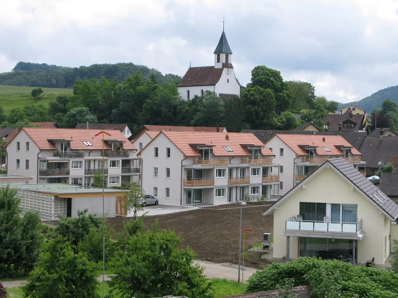Blick auf eine kleine Dorfszene mit modernen Mehrfamilienhäusern mit roten Dächern im Vordergrund, einer Kirche auf einem Hügel im Hintergrund, umgeben von Bäumen und grünen Hügeln.