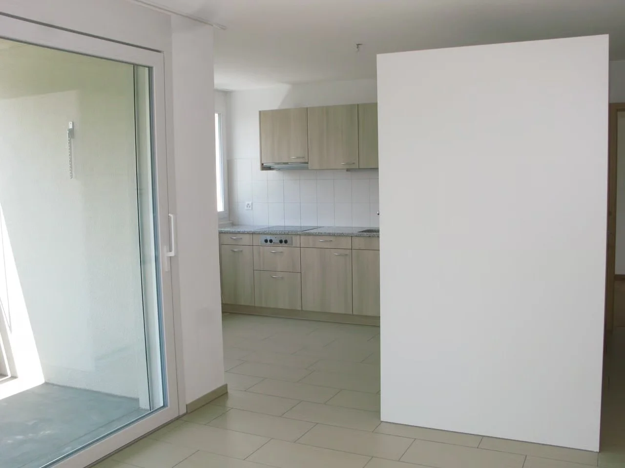 Leeres Kücheninterieur mit weißen und hellgrauen Schränken, einer Herdplatte und einer Fensterfront, in einem modernen Apartment