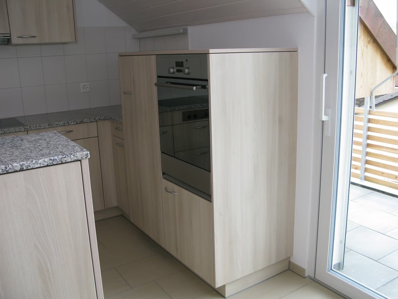 Küche mit Holzschränken, Granit-Arbeitsplatten, Backofen, Fliesenspiegel, auch ein Balkon mit Holzgeländer ist sichtbar