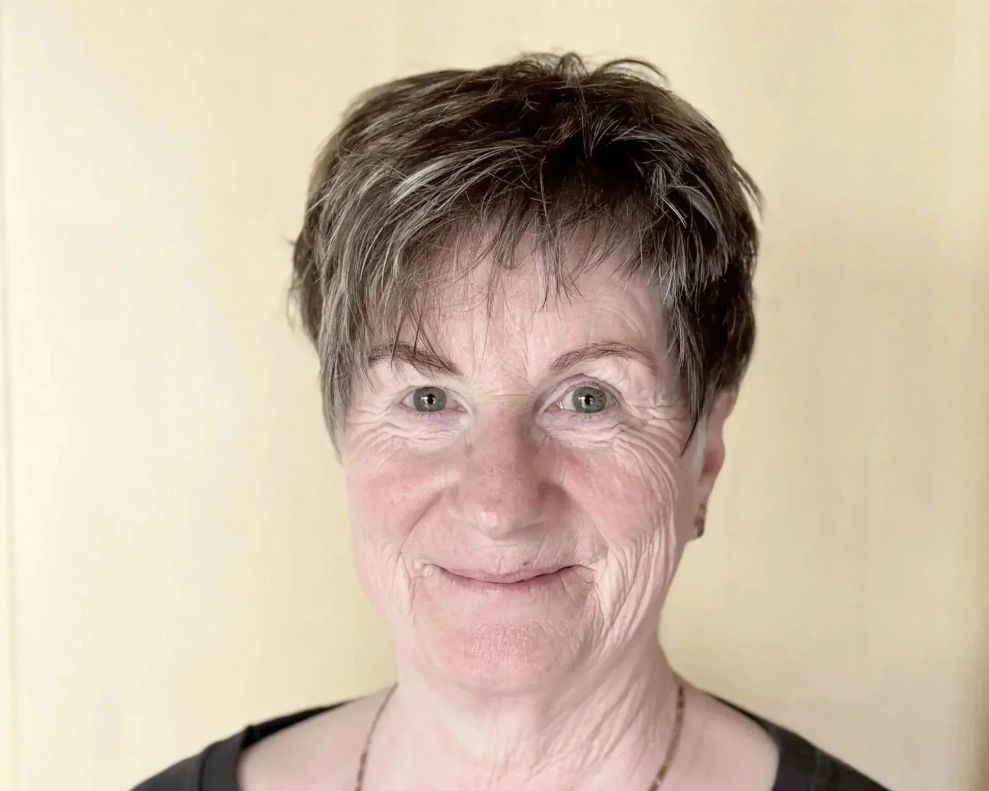 Nahaufnahme einer Frau mit kurze braune Haare, blauen Augen, einem schwarzen Oberteil und einem goldenen Halsketten vor einem hellen Hintergrund.