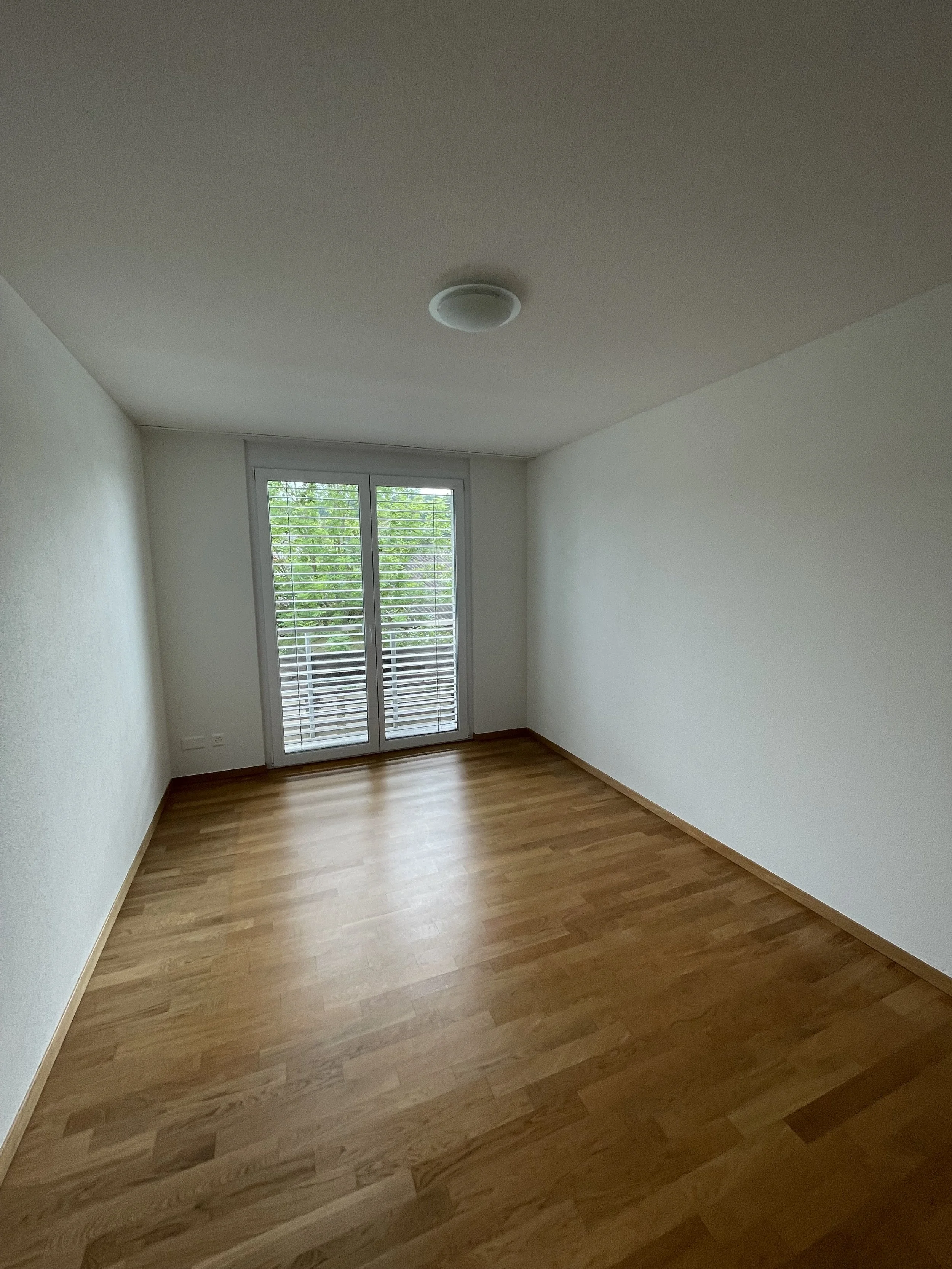 Leeres Zimmer mit Holzboden, weißen Wänden und einem Fenster mit Rollläden, das nach draußen blickt.