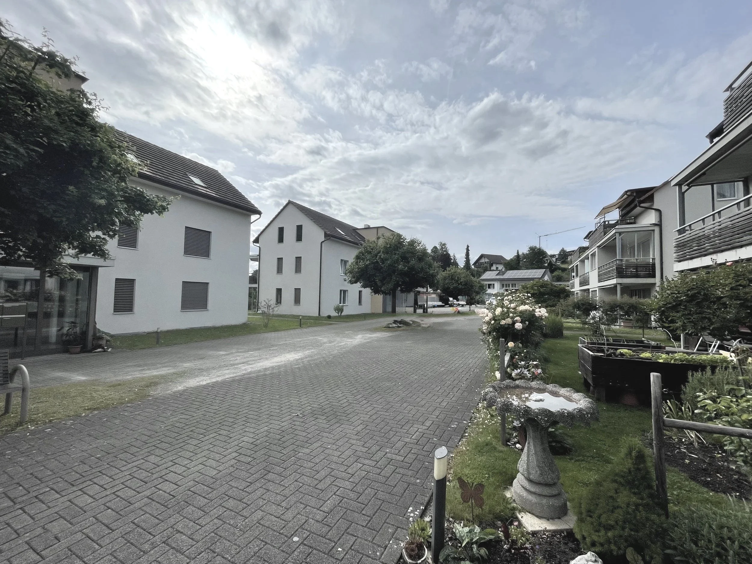 Ein ruhiger Wohnviertel mit weißen Mehrfamilienhäusern, einem gepflasterten Weg, Garten mit Blumen und Stauden, im Hintergrund bewölkter Himmel.