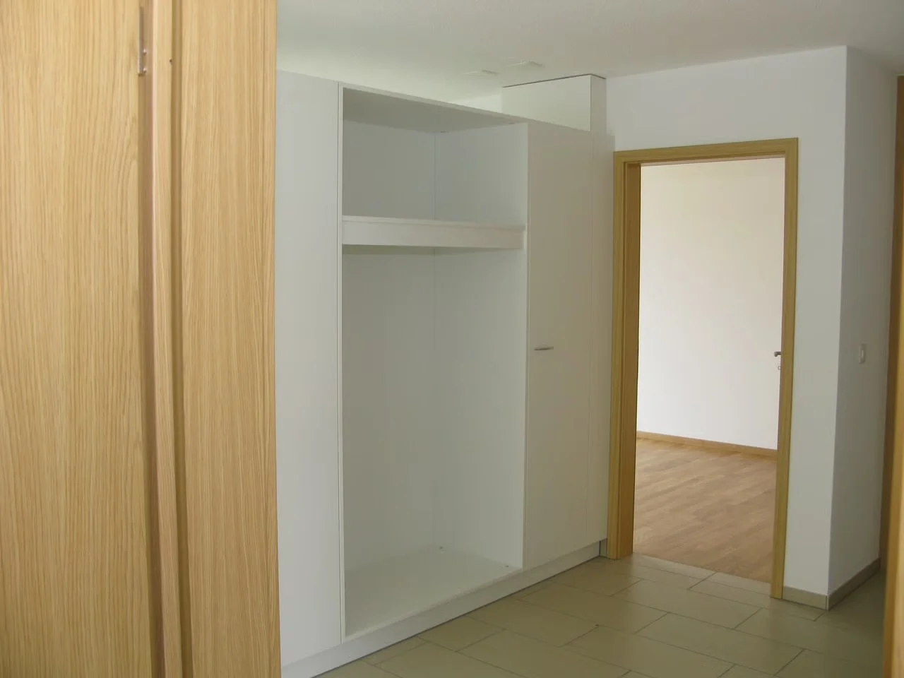 Leere Einbauschrank mit weißem Innenraum in einem Flur, daneben eine Holztür zum angrenzenden Raum mit Holzboden.