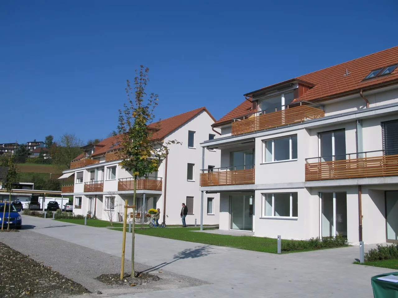 Mehrstöckiges modernes Wohnhaus mit weißen Wänden, Holzbalkonen, roten Ziegeldächern und kleinen Gärten im Vordergrund, sonniger Himmel.
