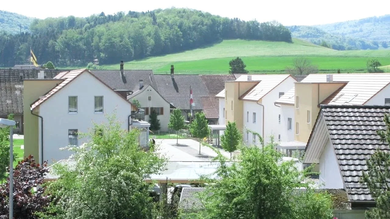 Blick auf Gässlimat in Bözen mit weißen und hellgelben Häusern, umgeben von grünen Bäumen, mit einer grünen Hügellandschaft im Hintergrund.