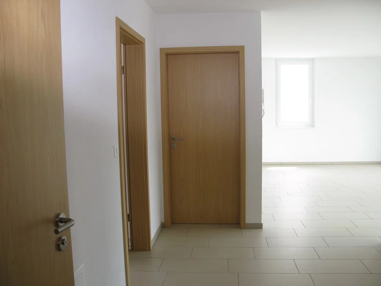 Ein modernes, leerstehendes Zimmer mit weißen Wänden, Holztüren und großen Fenstern, hell erleuchtet durch Tageslicht.