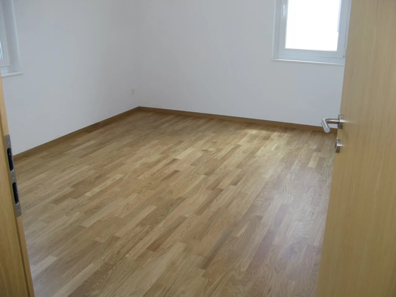 Leeres Zimmer mit Holzfußboden und weißen Wänden, Fenster auf der rechten Seite, offenes Tür.