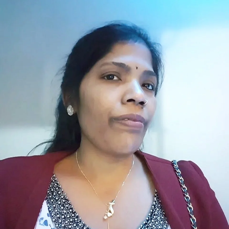 Nahaufnahme einer Frau mit dunklem Haar, goldener Halskette, Ohrringen und Nasenpiercing, trägt eine rote Jacke und ein schwarz-weiß gemustertes Oberteil, sitzt vor einem hellen Hintergrund.