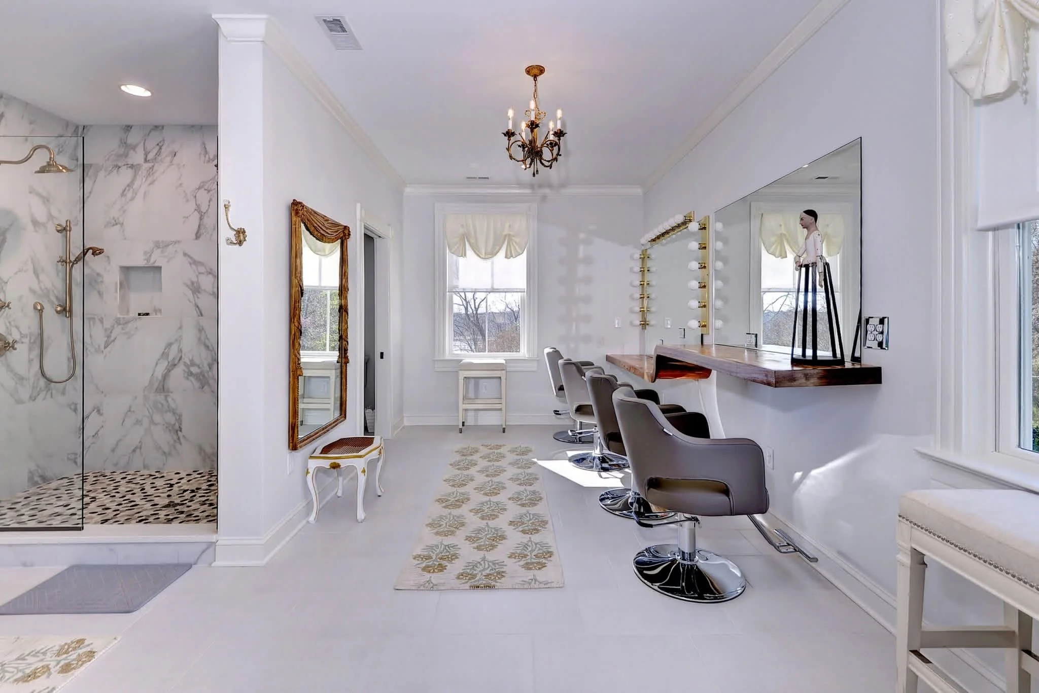 An elegant bride's dress and bouquet placed on a beige armchair with a pair of nude high heel shoes on a wooden floor, in a bright room with white walls, a white vanity table, and a mirror.