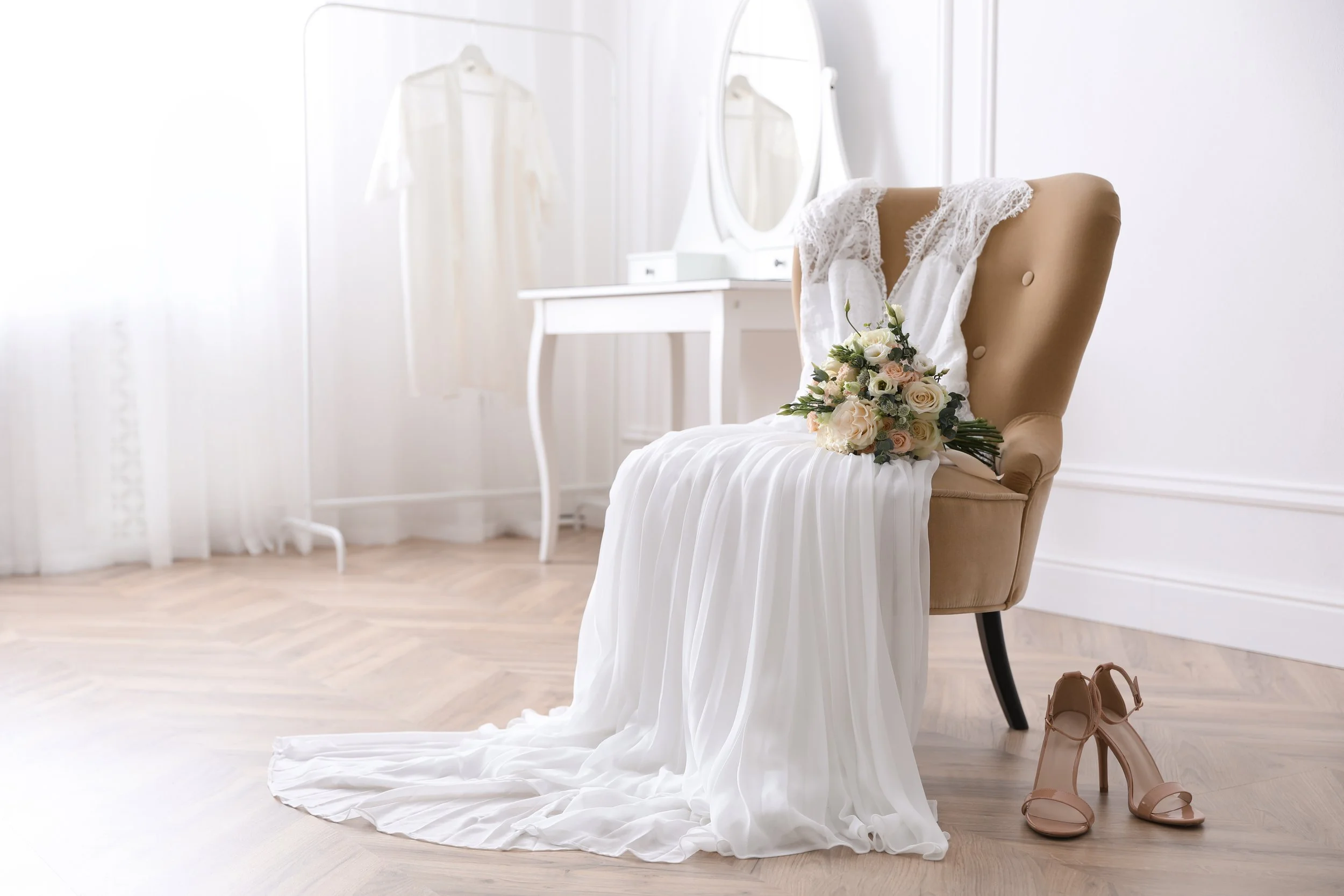 An elegant bride's dress and bouquet placed on a beige armchair with a pair of nude high heel shoes on a wooden floor, in a bright room with white walls, a white vanity table, and a mirror.