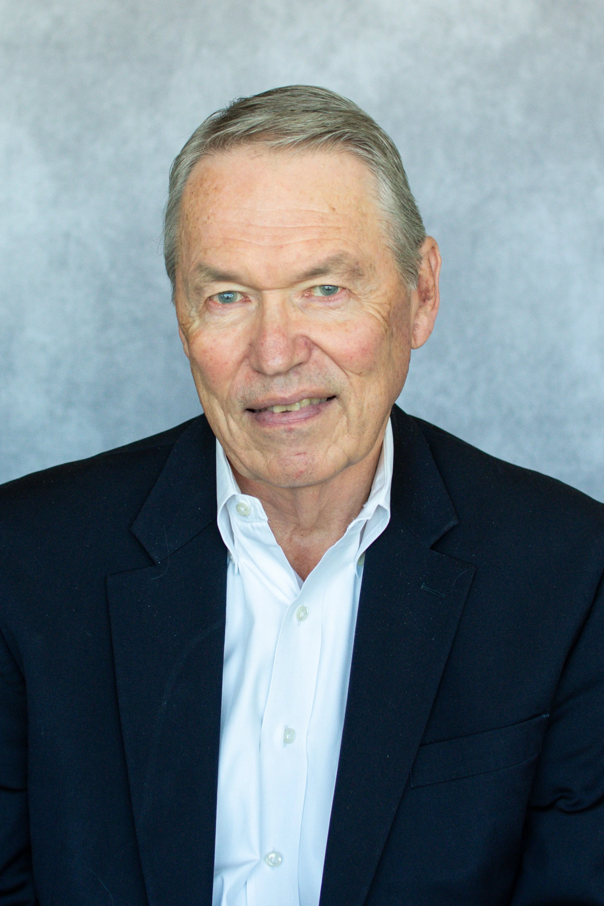 A mature man with gray hair wearing a dark suit jacket and a white collared shirt, smiling at the camera, against a neutral gray background.