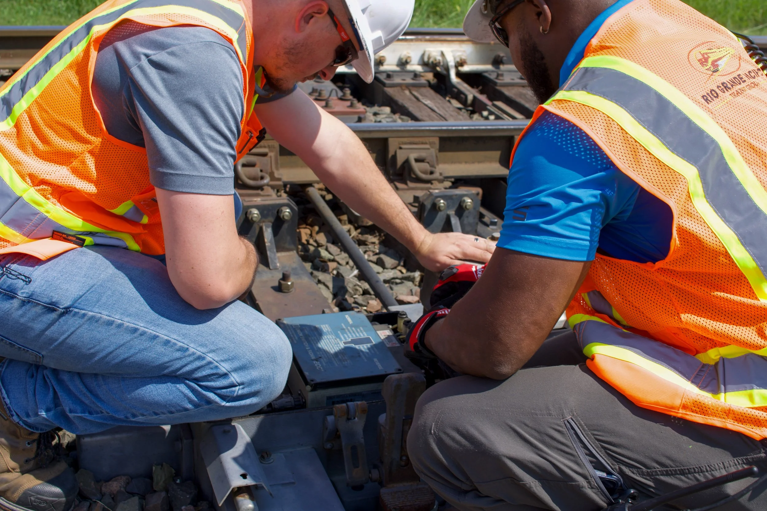 Two railway workers in safety vests and hats inspecting and repairing a section of train tracks outdoors.