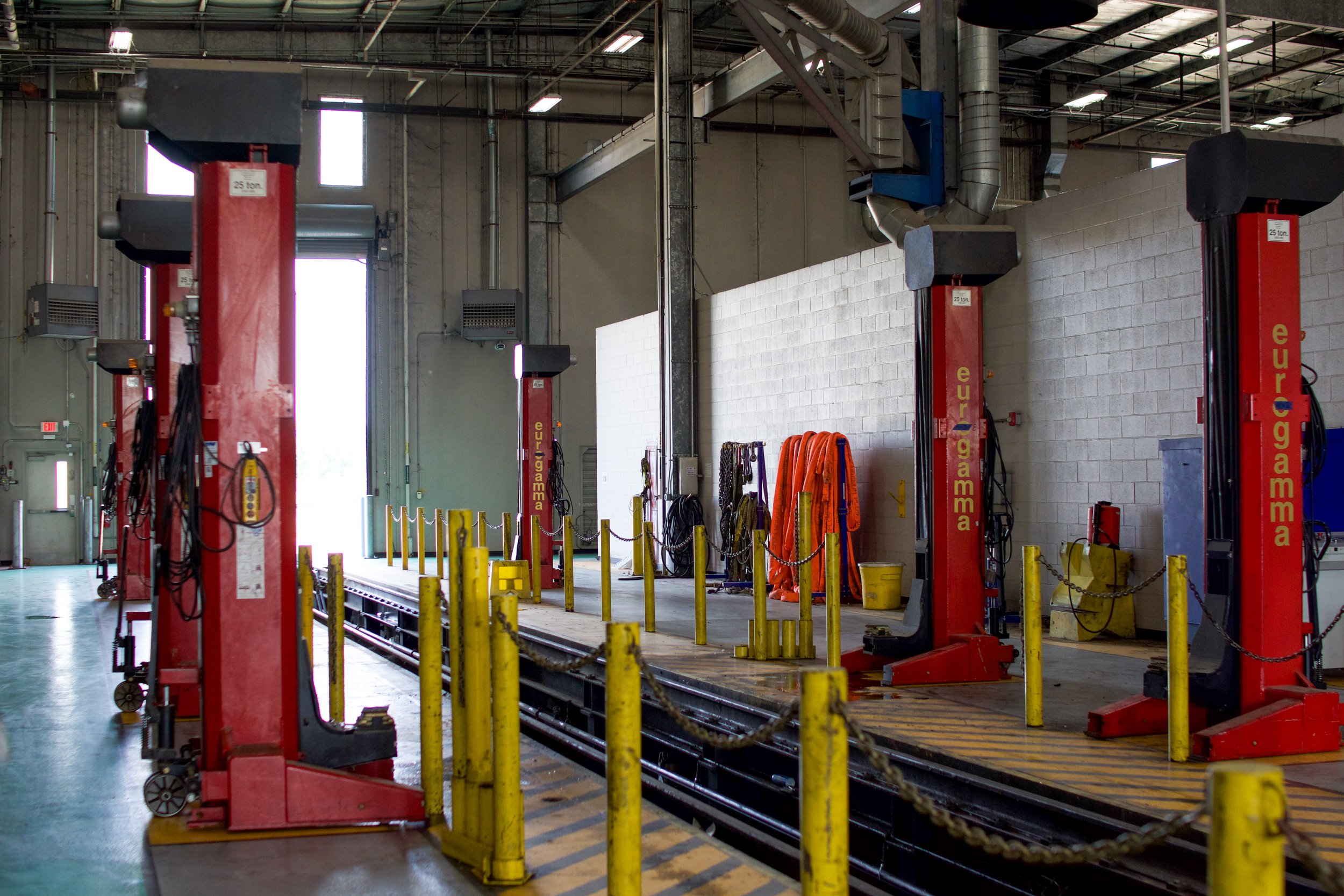 Industrial work area with red lifts labeled 'euro gamma', yellow safety posts and chains, various tools and orange safety vests hanging on a wall, in a spacious warehouse with high ceilings and PVC pipes.