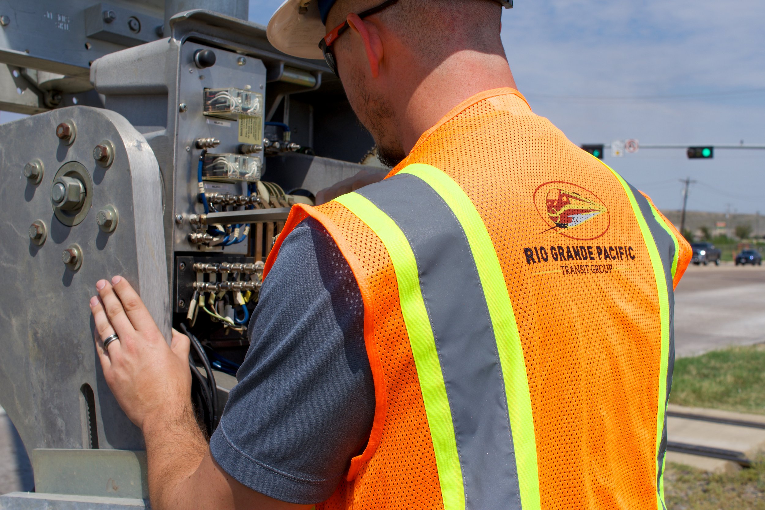 A worker wearing a hard hat and safety vest working on electrical equipment on a city street with traffic lights and cars in the background.