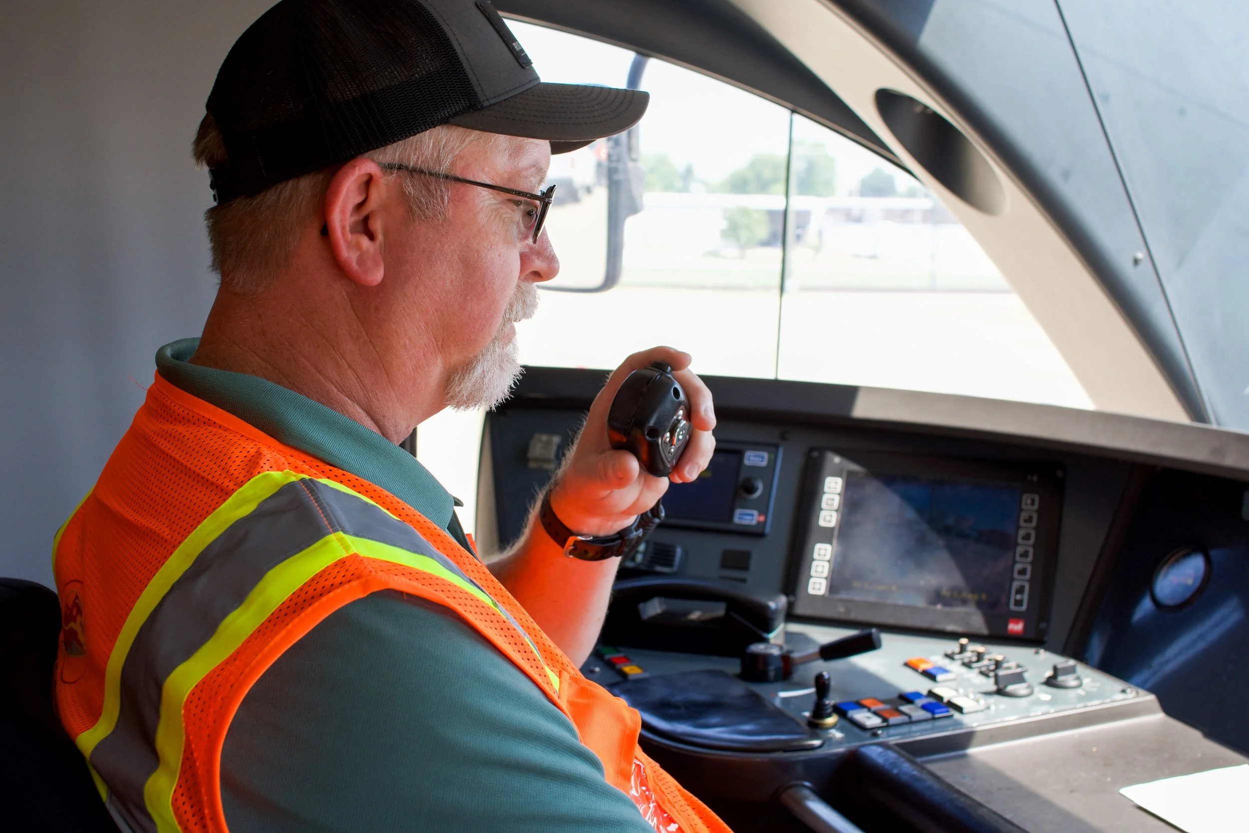 An older man with a white beard, glasses, wearing a black cap and an orange safety vest, is sitting in the cockpit of a train or tram, holding a control device, with a digital display and controls in front of him visible.